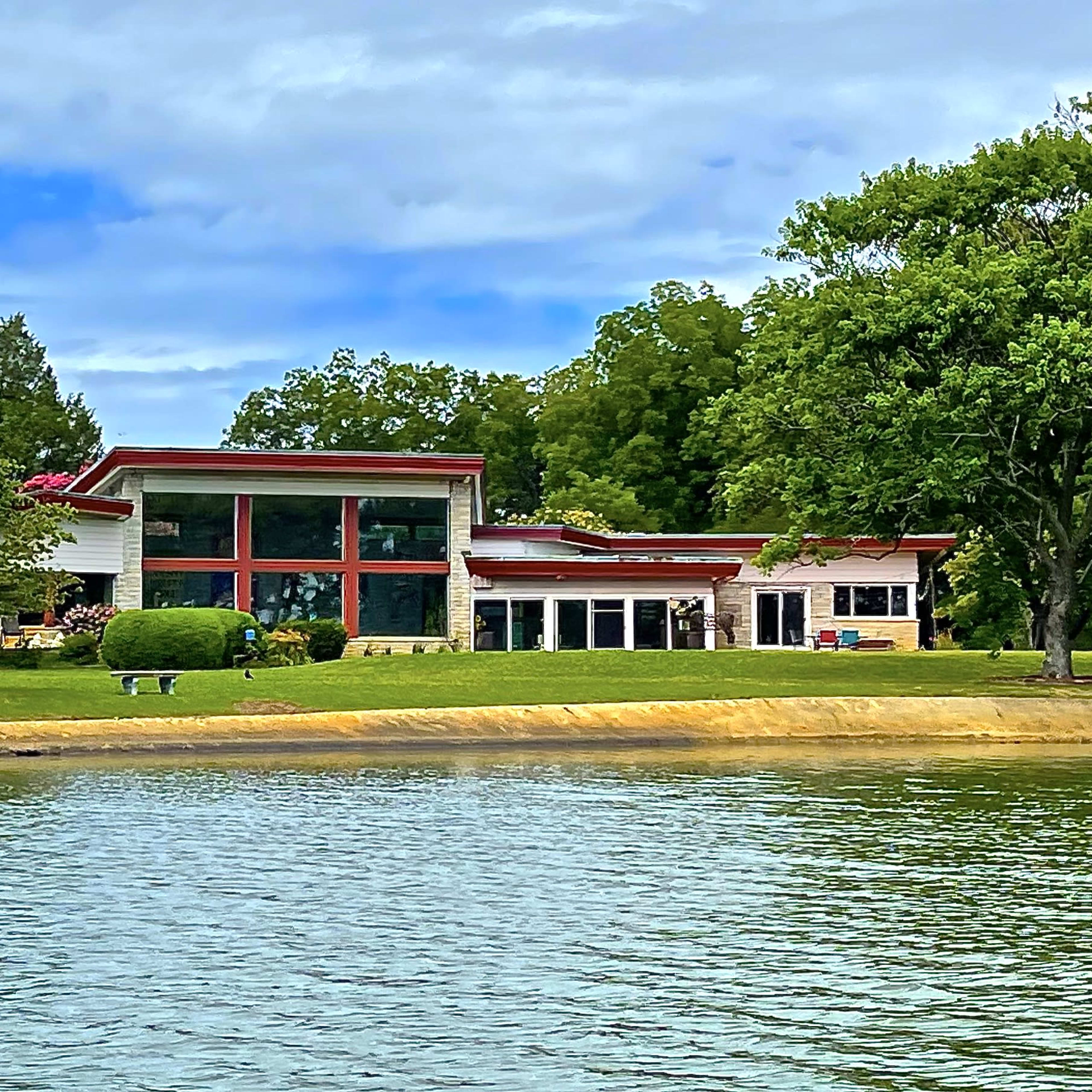 A modern house with large windows sits beside a calm lake, surrounded by trees and well-manicured lawns.