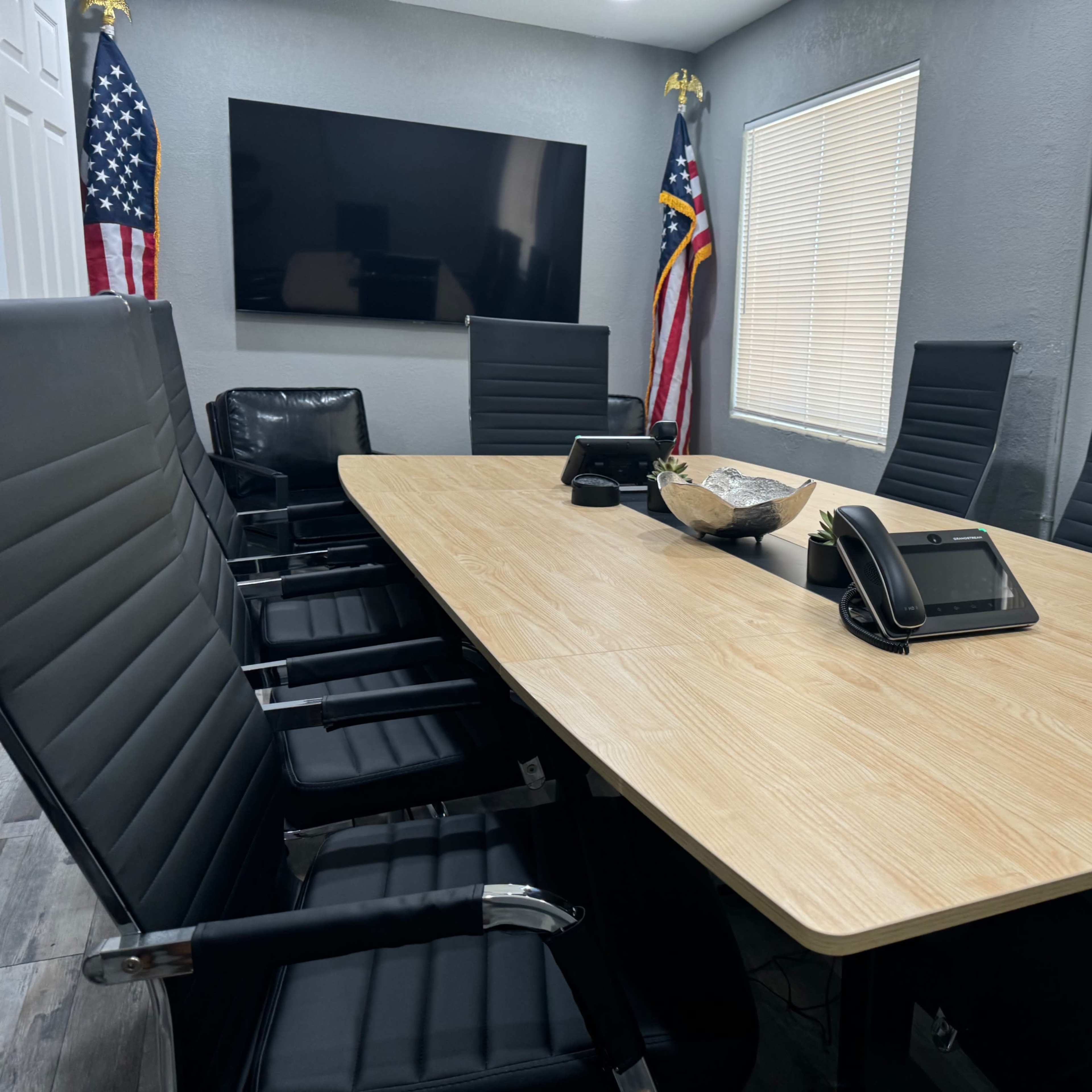 A large conference room features a wooden table surrounded by several black chairs, with a television on the wall and American flags in the background.