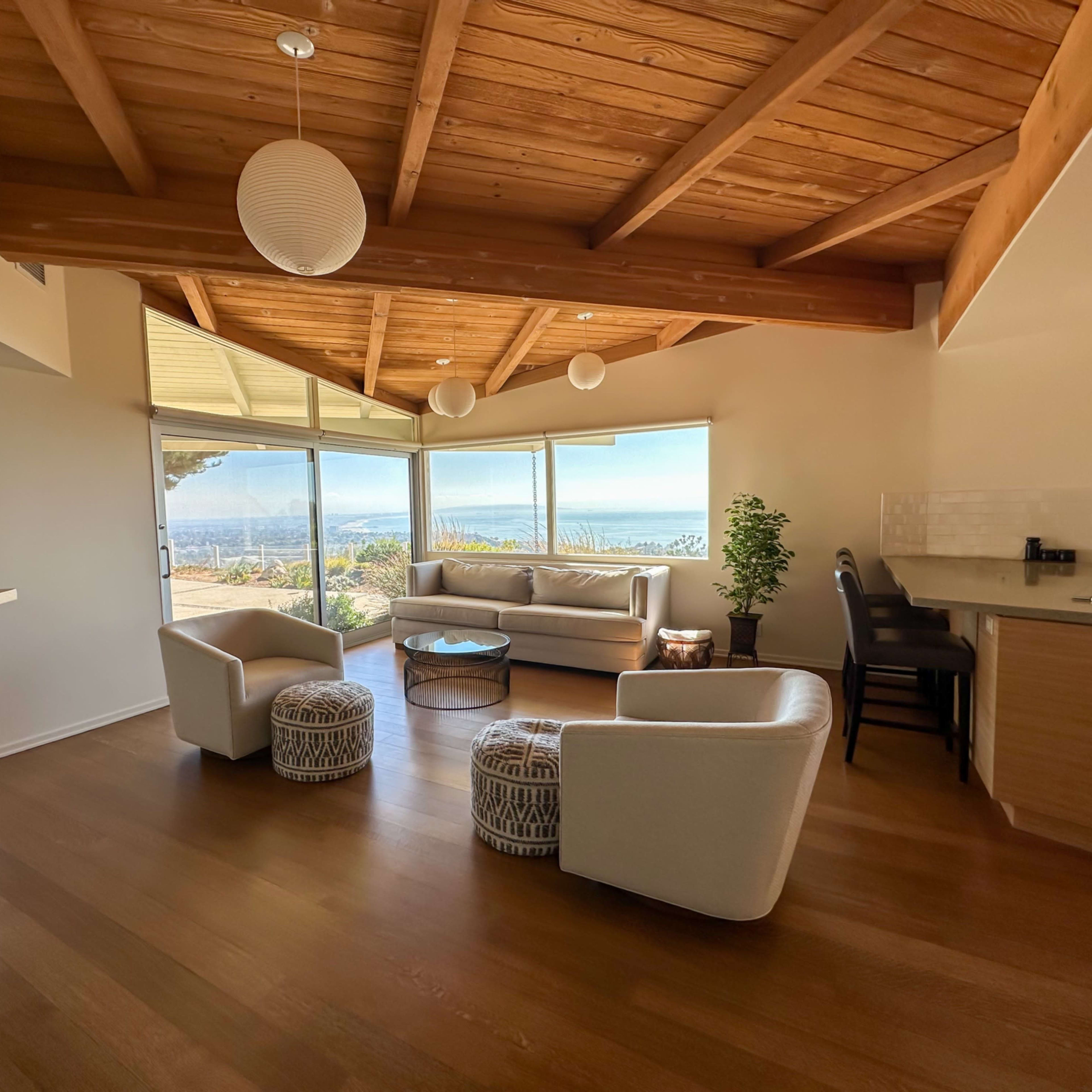 A modern living room with large windows overlooking a scenic view, featuring a beige sofa, two armchairs, a coffee table, and wooden beams on the ceiling.