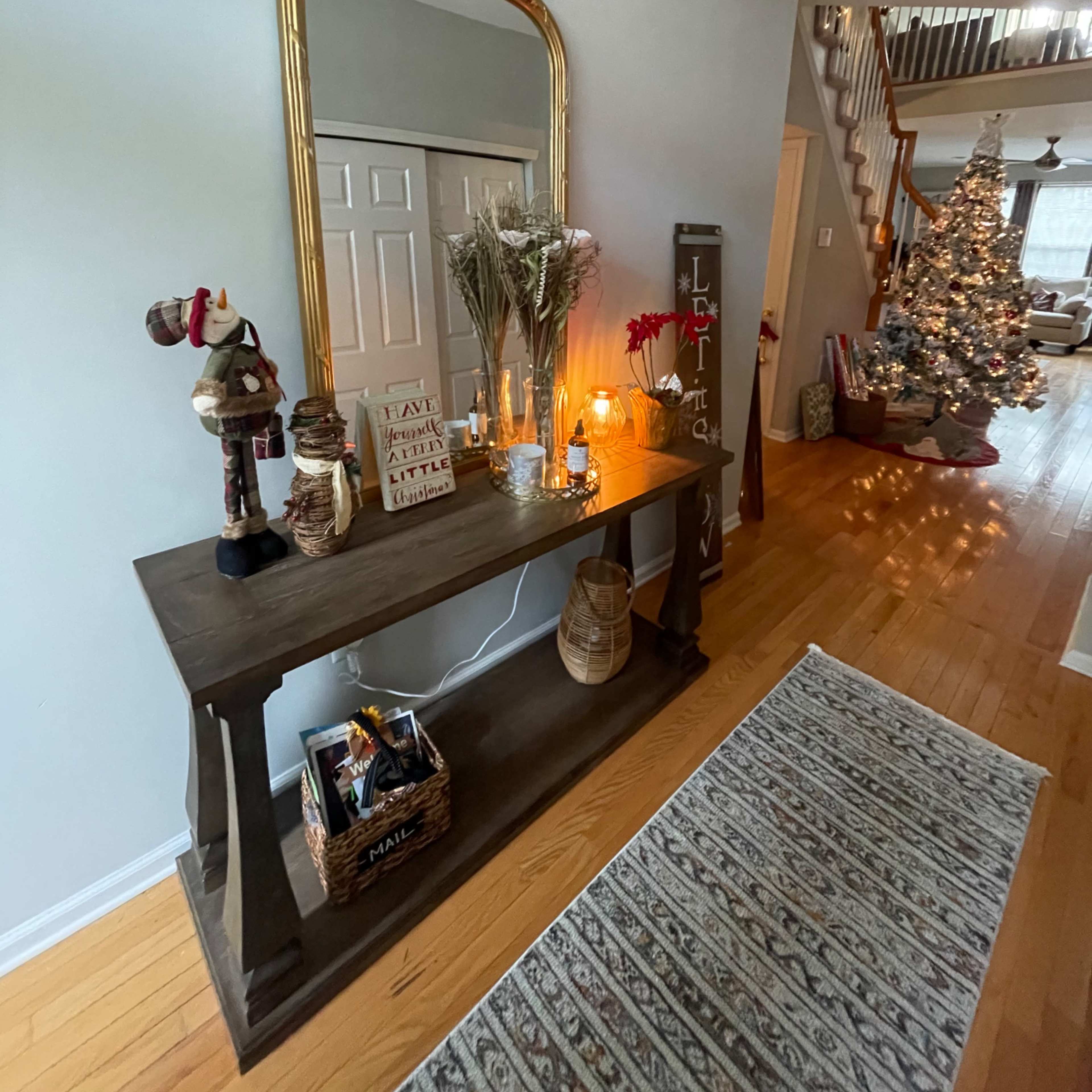 A console table adorned with decorative items, a large mirror, and candles is positioned against a light-colored wall in a hallway, with a Christmas tree visible in the background.