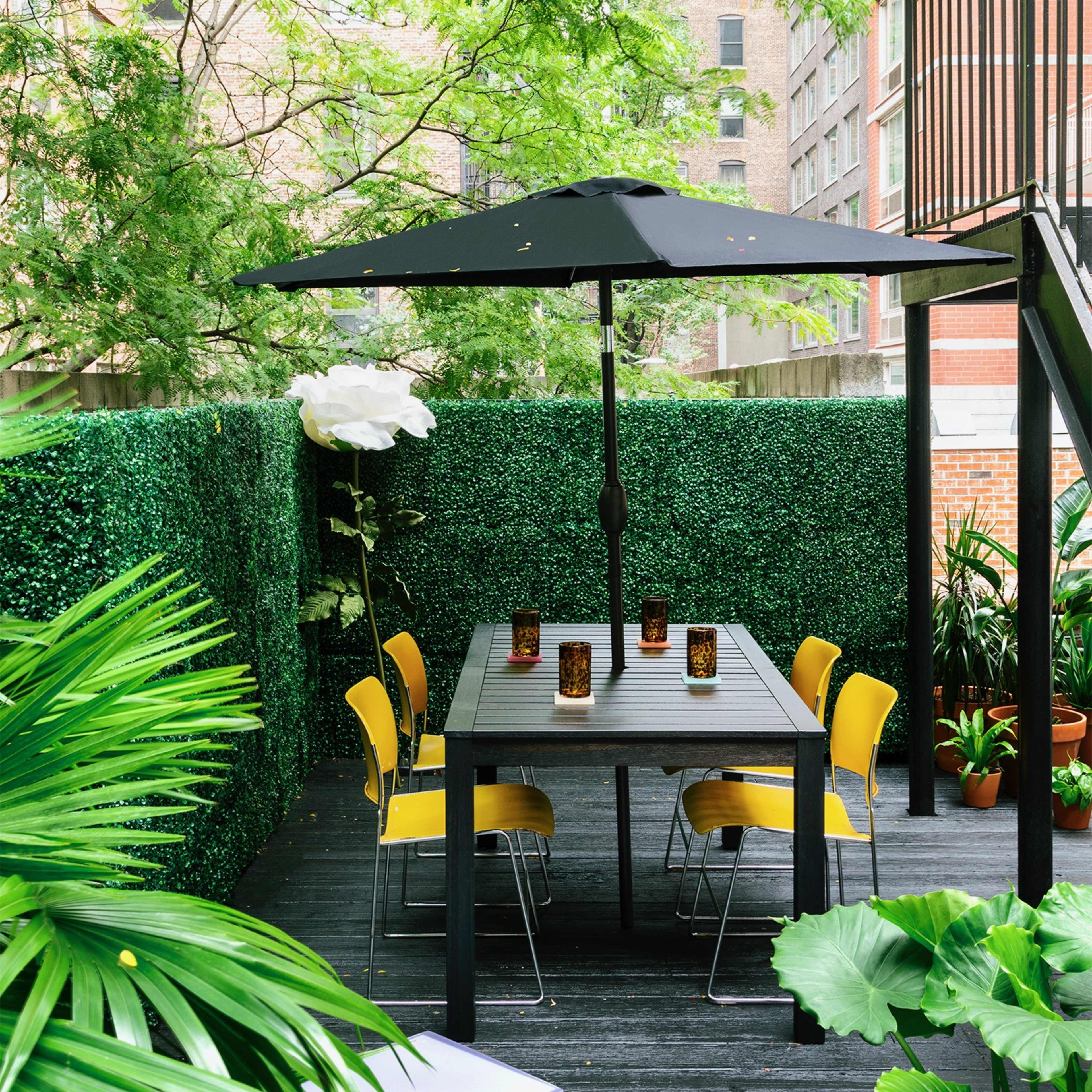 A dining table with yellow chairs is set under a black umbrella in a green, landscaped outdoor space surrounded by tall plants and a hedge.