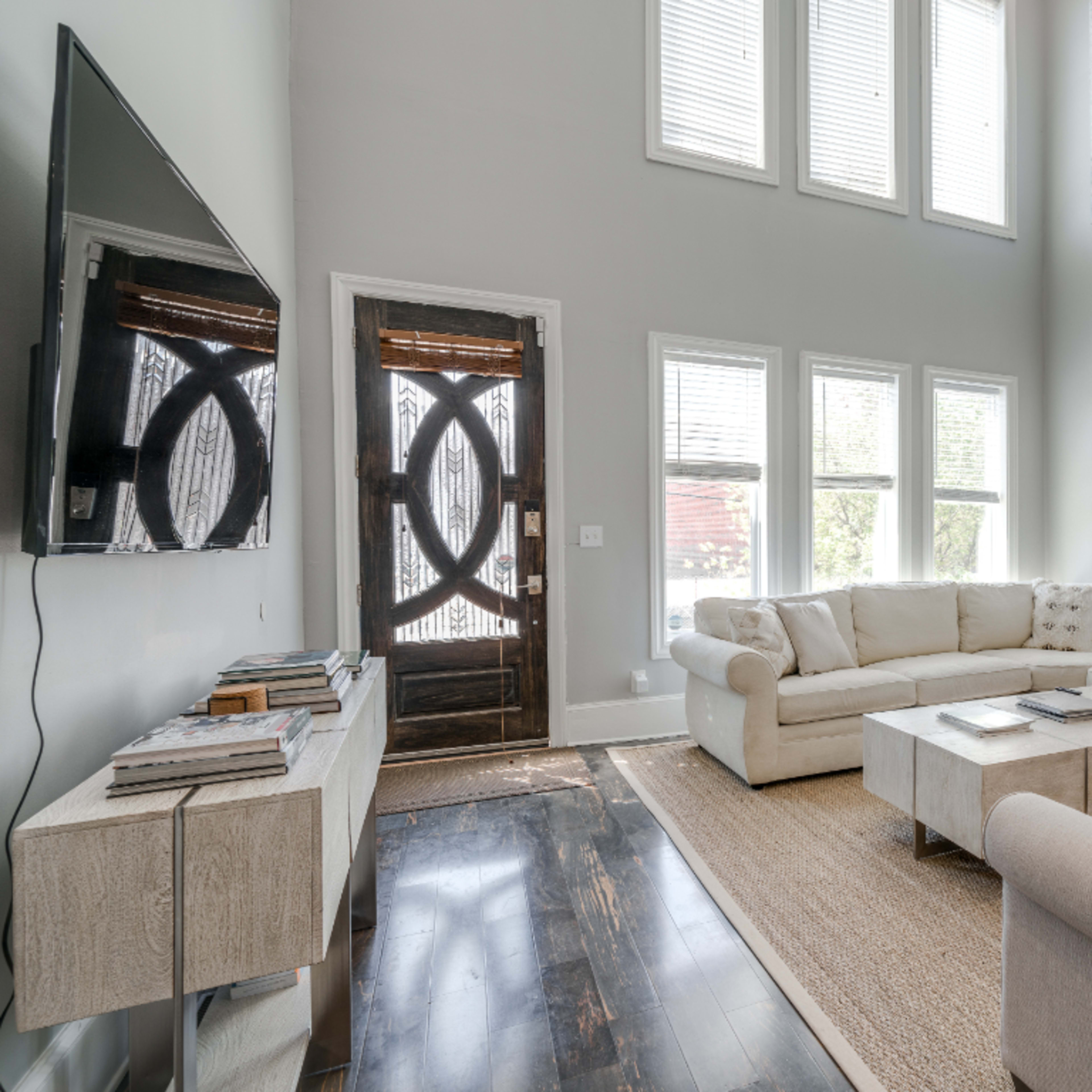 A modern living room with a large sectional couch, a coffee table, a flat-screen TV mounted on the wall, and bright windows allowing natural light.