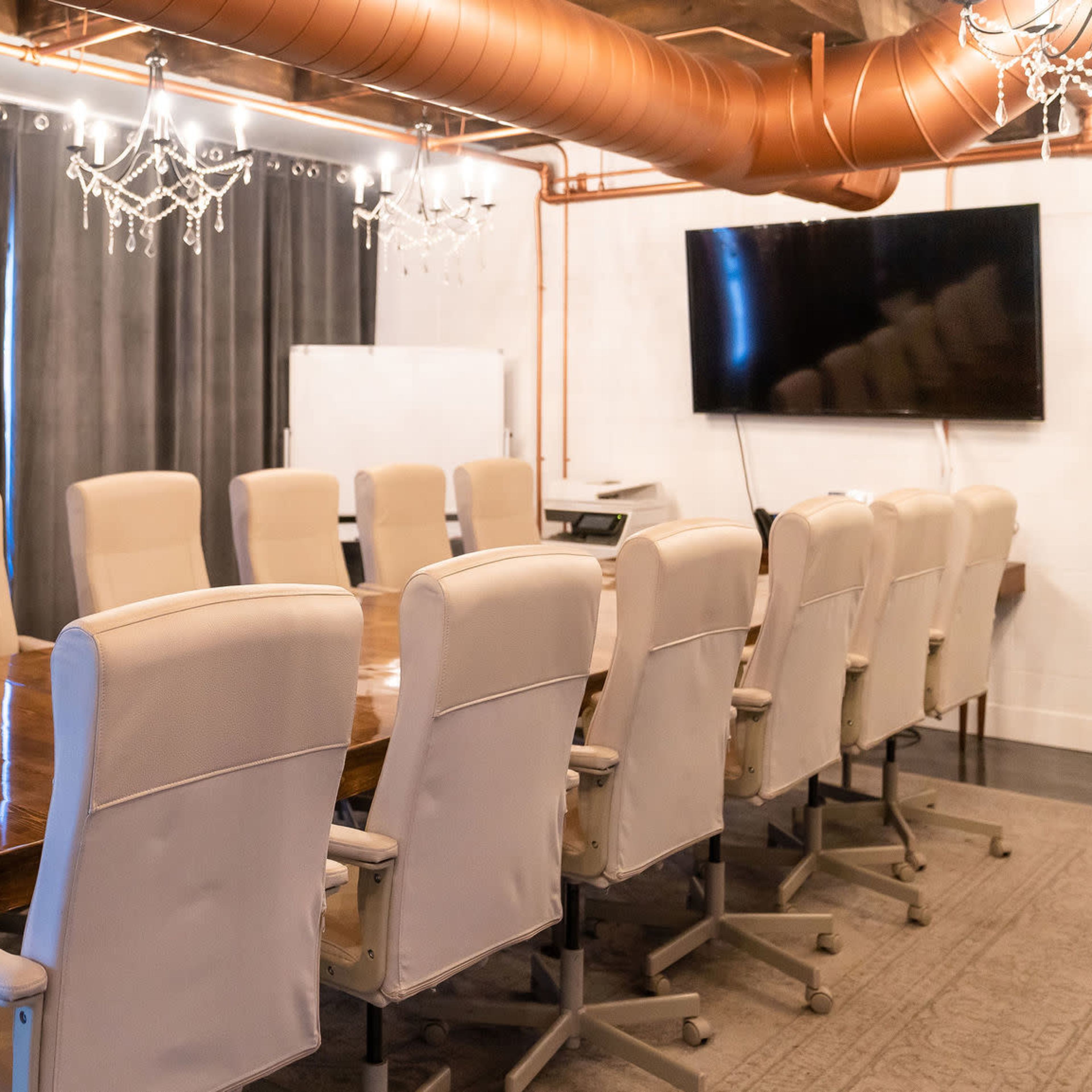 A spacious conference room features a large wooden table surrounded by twelve beige chairs, with curtains and chandeliers enhancing the decor.