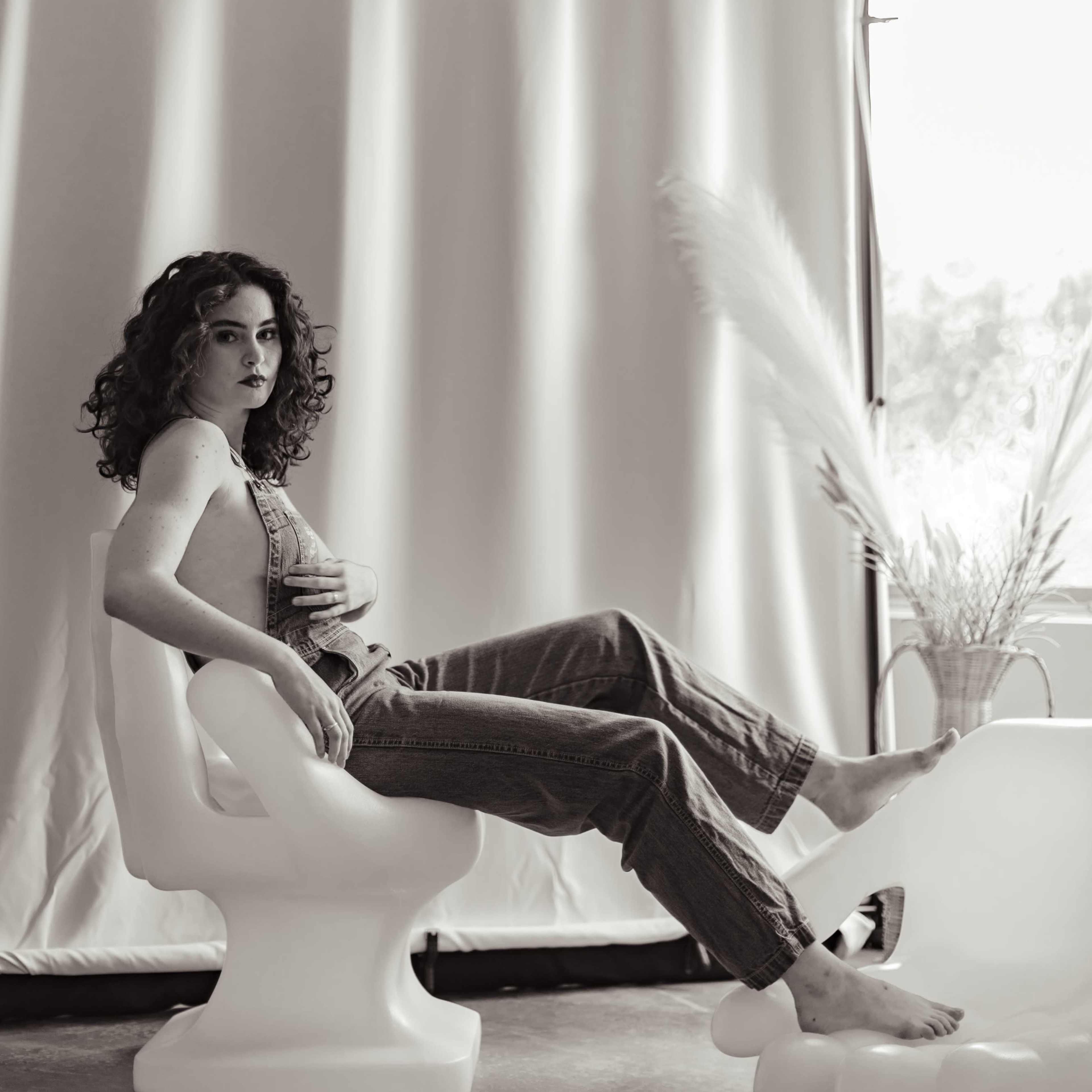 A person with curly hair sits on a modern chair in a well-lit room, with a curtain in the background and a plant nearby.