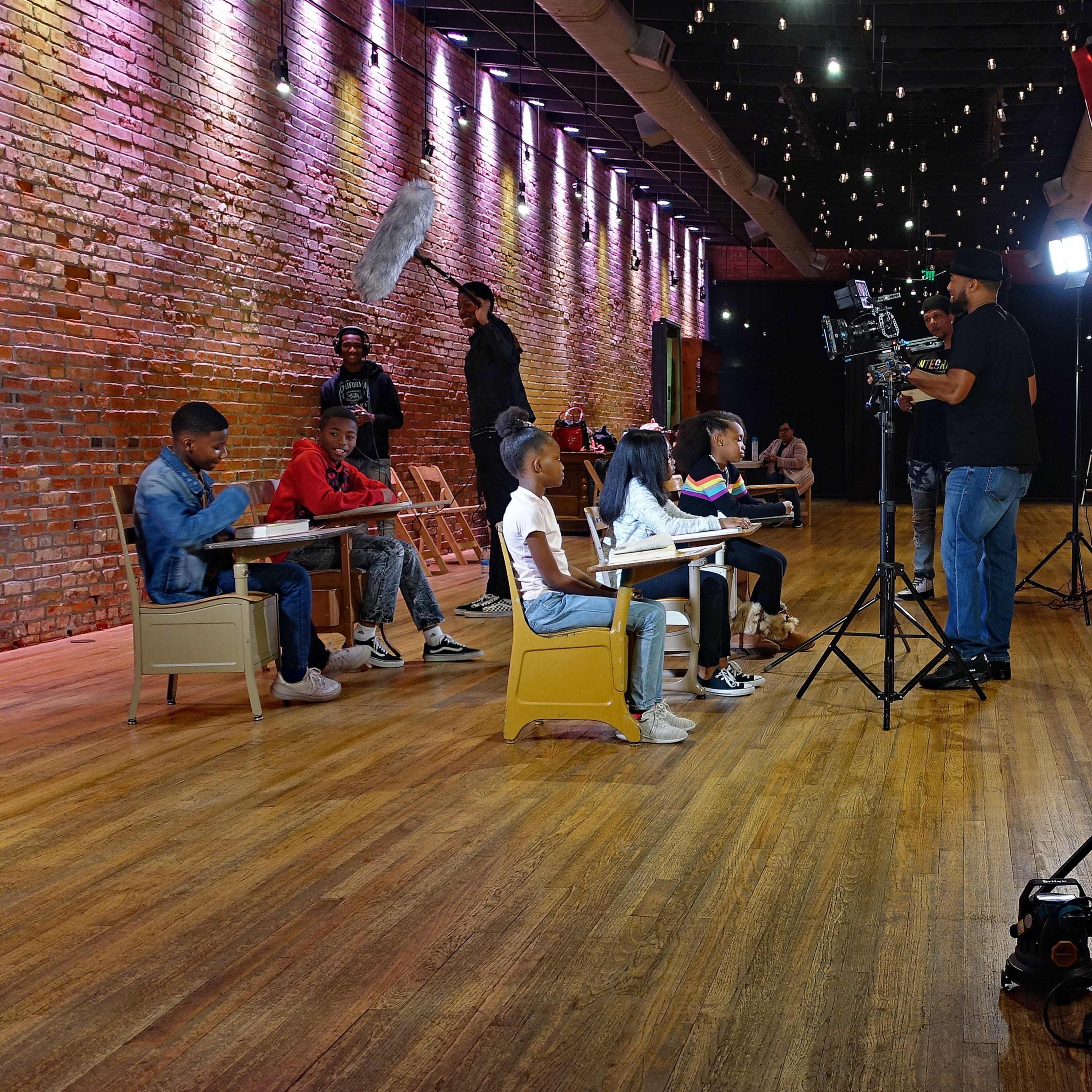 A classroom setting with students seated at desks while a crew films a scene with cameras and lighting equipment.