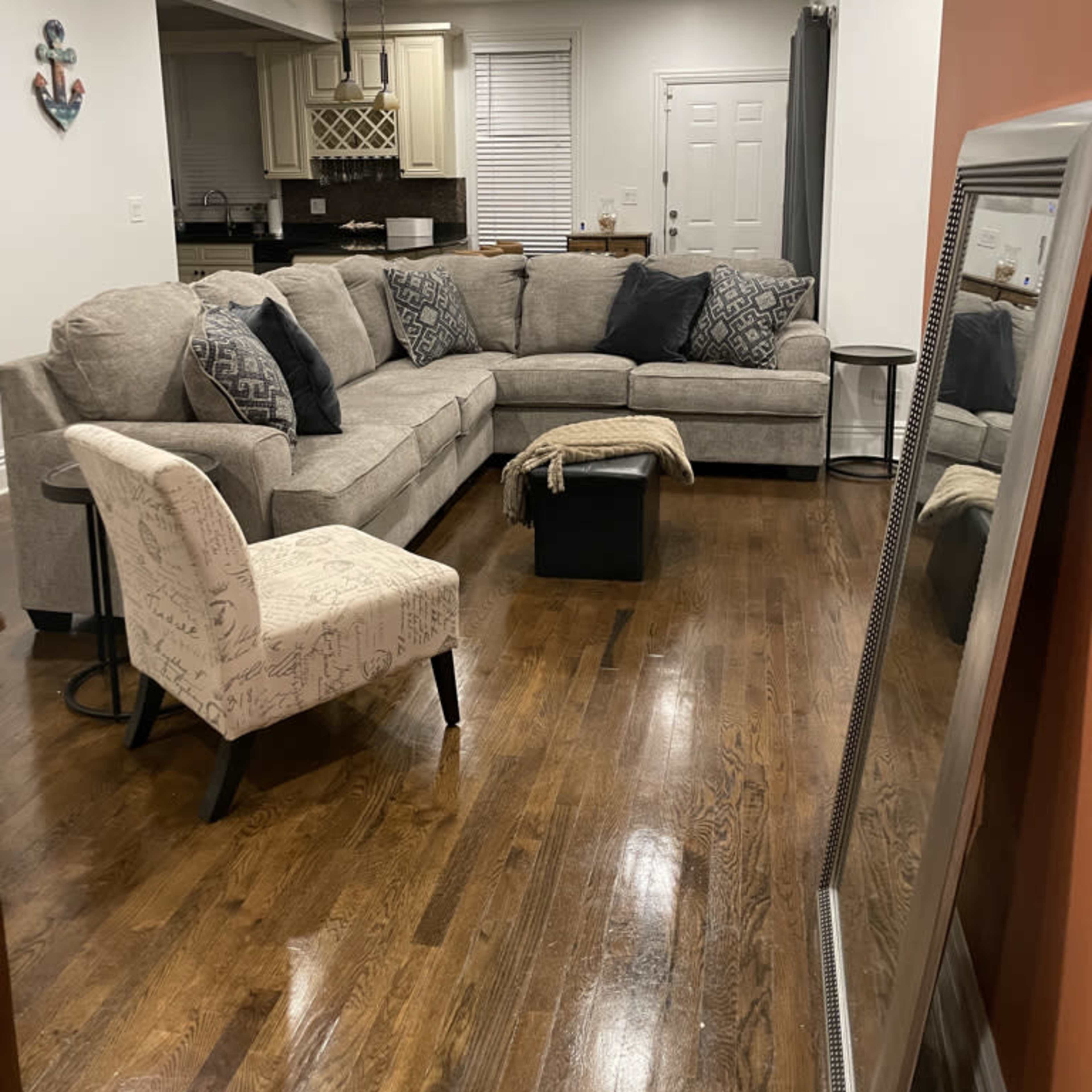 A cozy living room features a large sectional couch, an accent chair, a coffee table, and a mirror, with hardwood flooring and a view into a kitchen area.