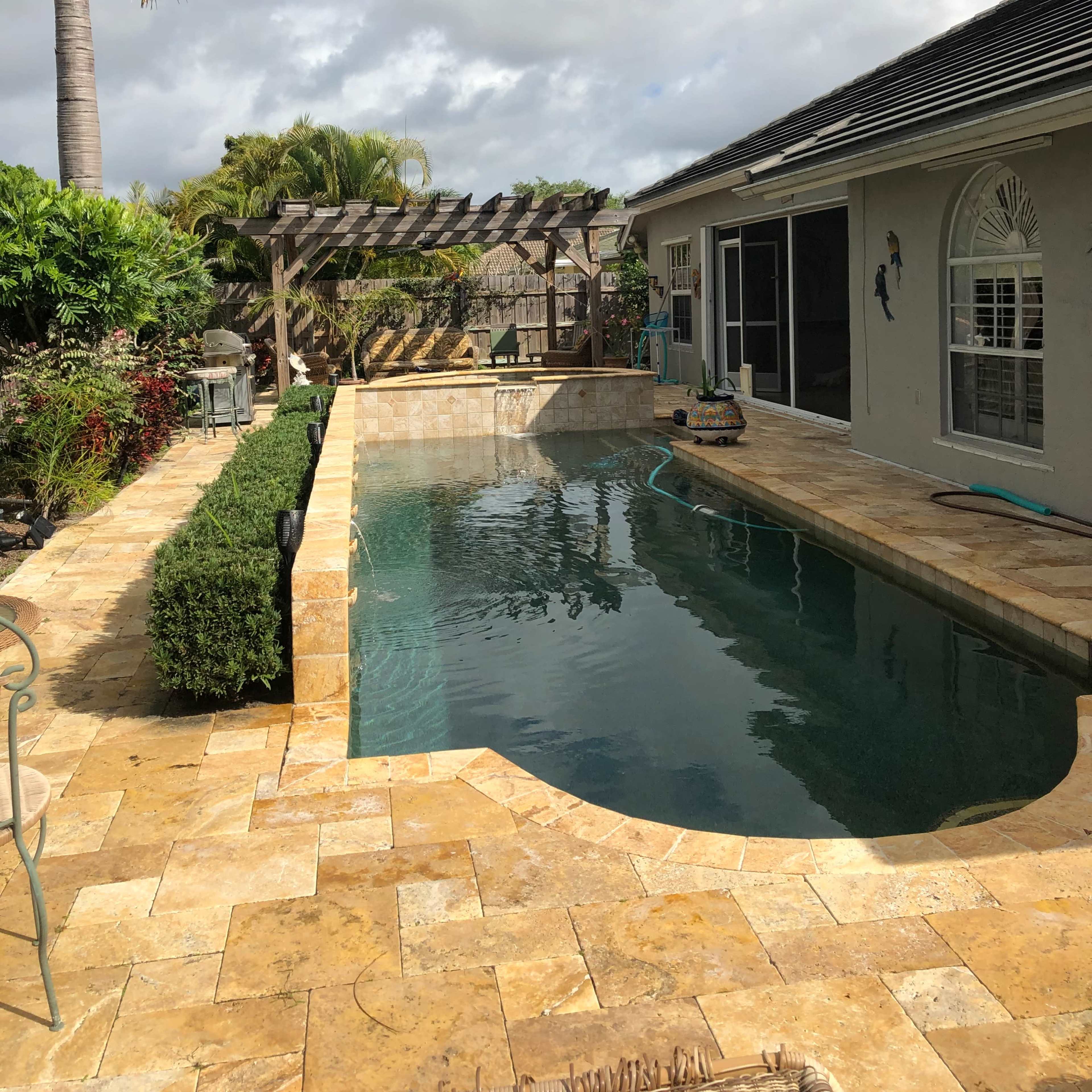 A backyard swimming pool area with stone tiles, surrounded by greenery and a wooden pergola, under a partly cloudy sky.