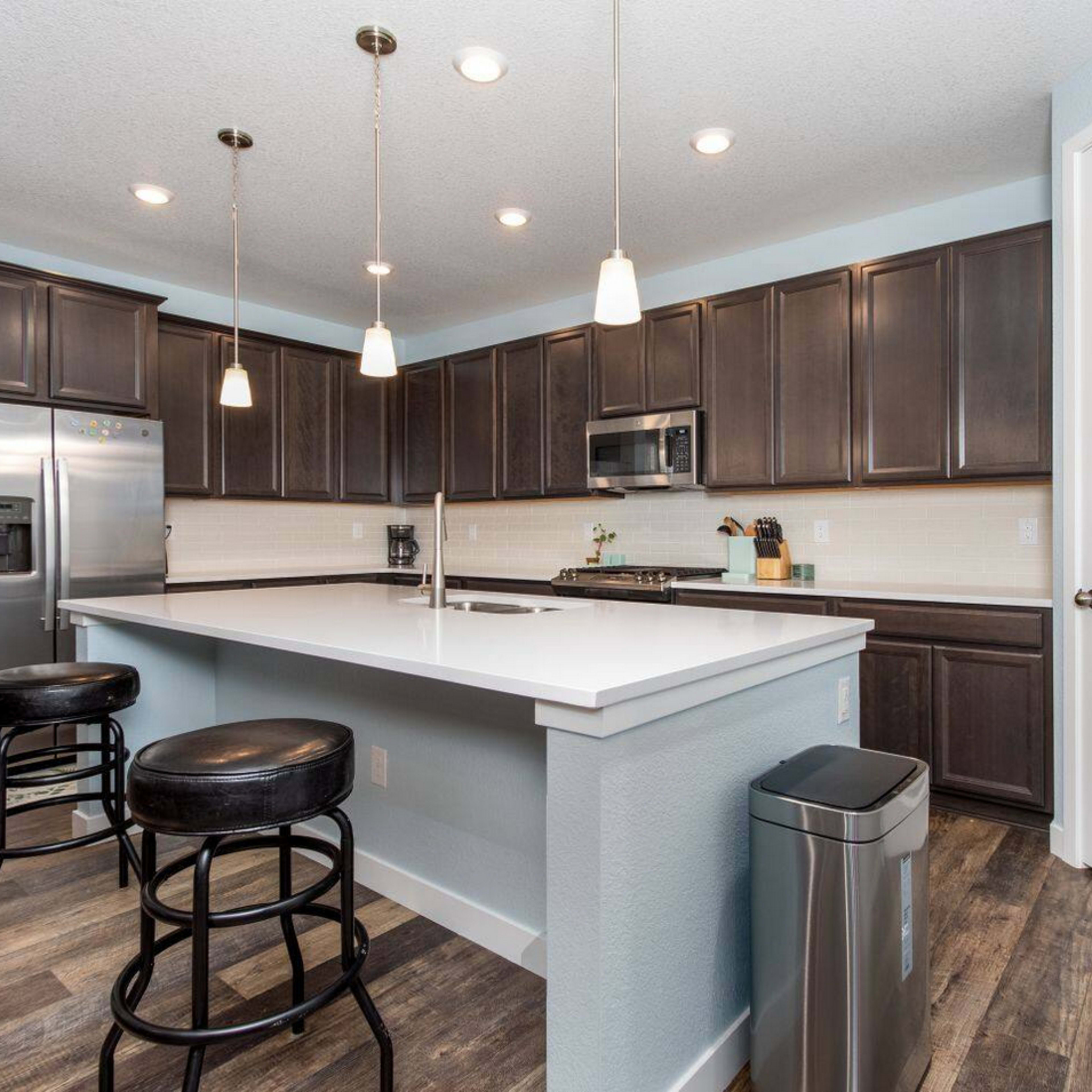 A modern kitchen features dark wood cabinets, a white countertop with bar seating, stainless steel appliances, and pendant lighting.