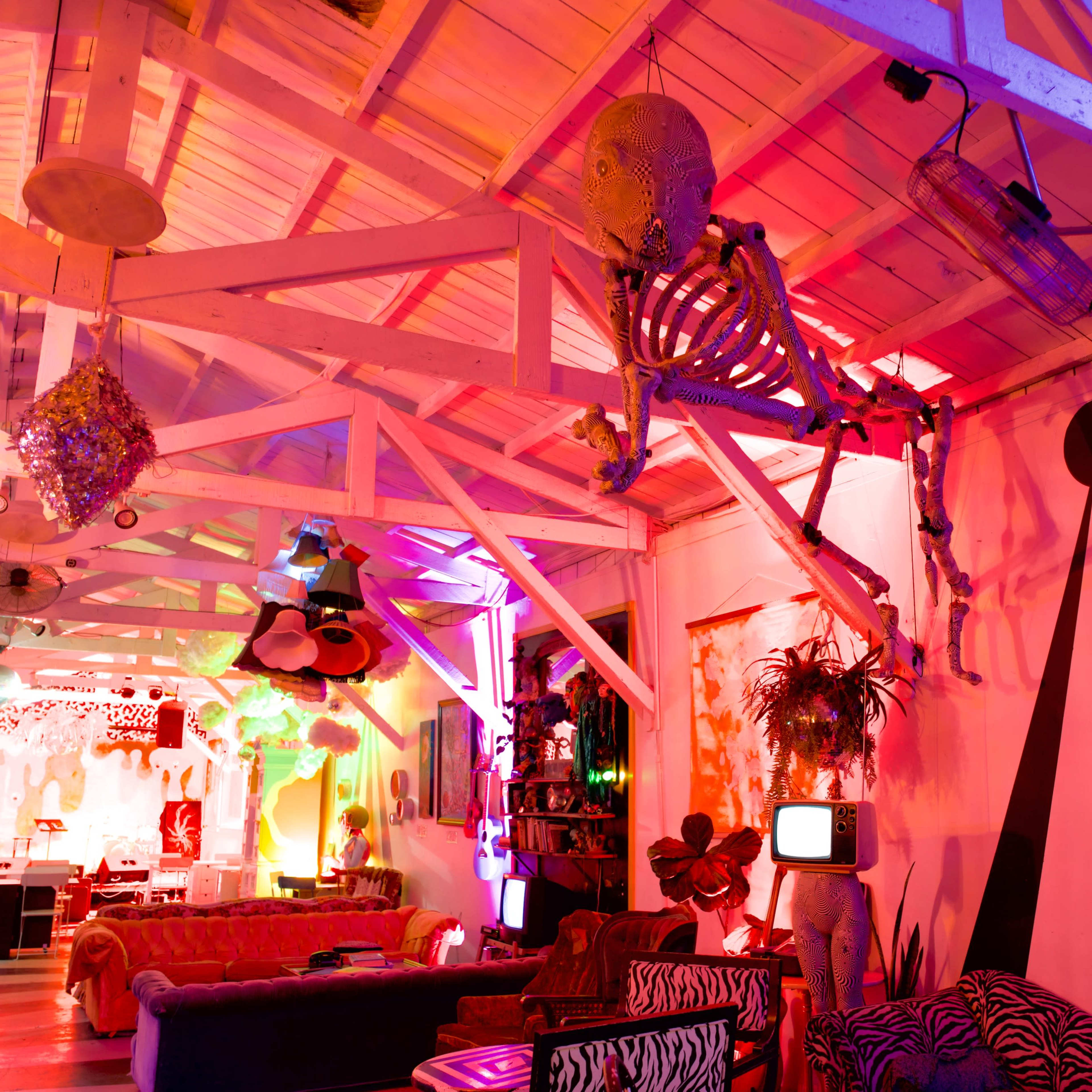 The interior of a colorful, artistic lounge features a skeleton suspended from the ceiling, surrounded by various eclectic decorations and vibrant lighting.