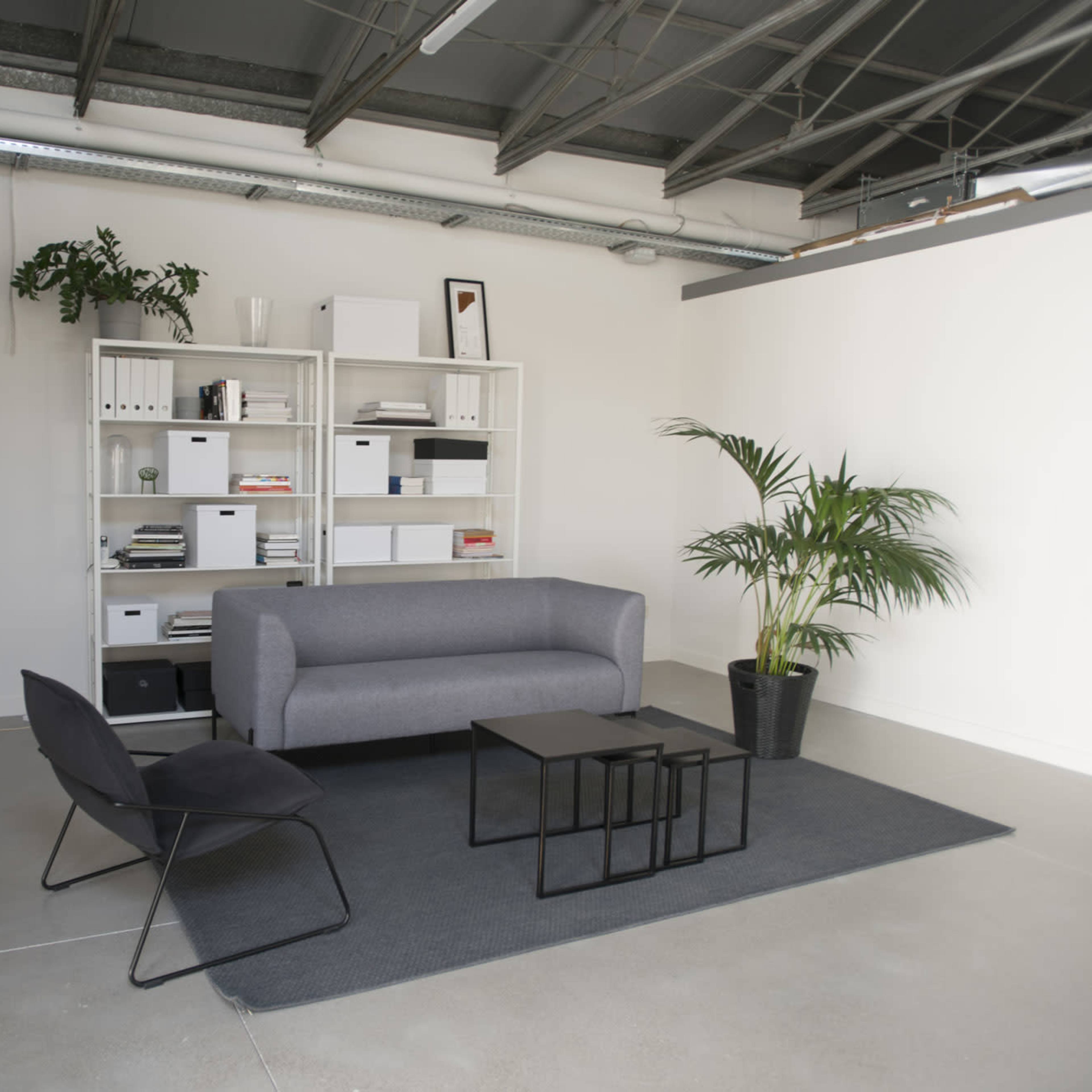 A minimalist office space features a gray couch, a black chair, a black coffee table, a light gray rug, and a shelving unit with storage boxes against a white wall.