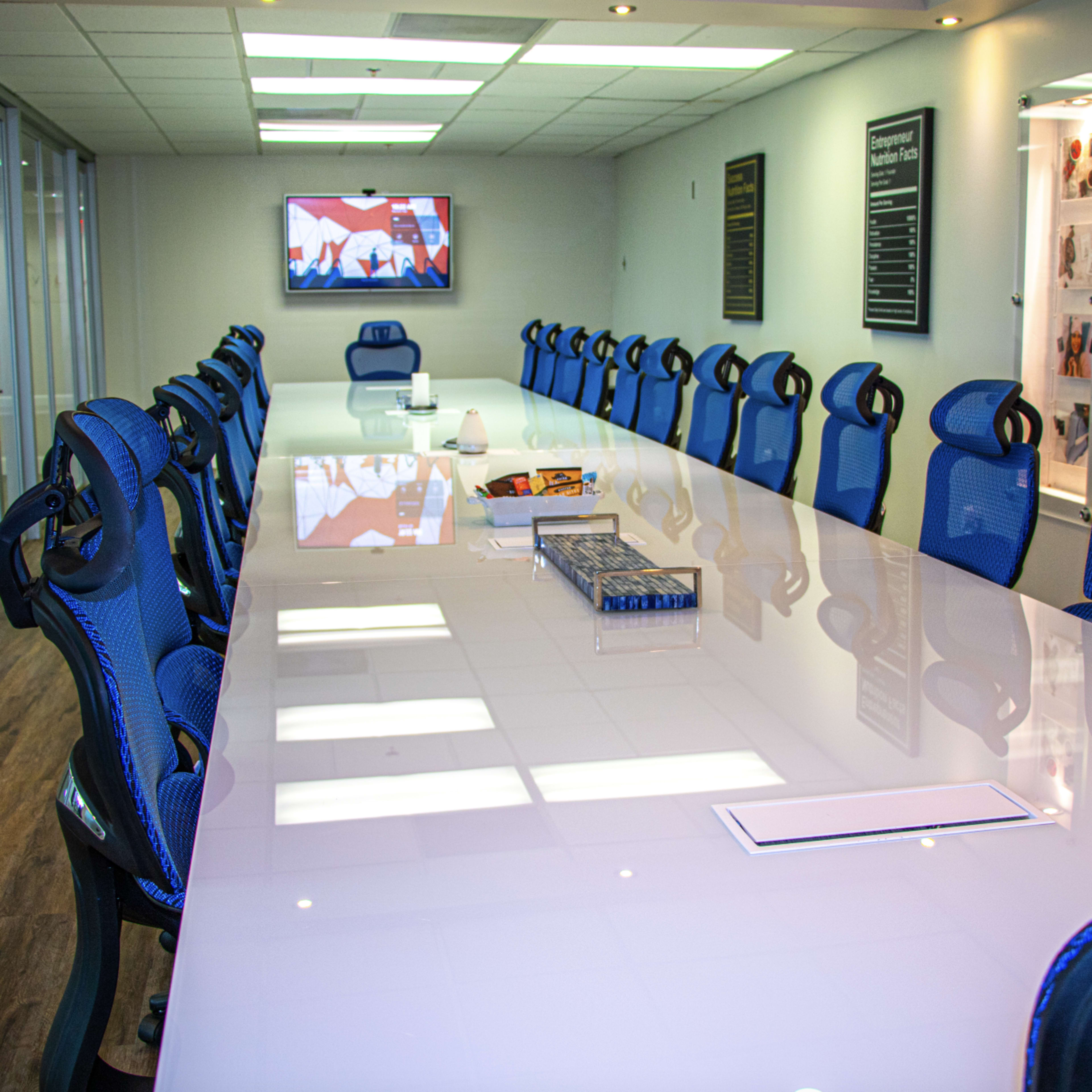 A long, modern conference table with blue chairs is set up in a brightly lit meeting room, featuring a television on the wall and a display case in the background.
