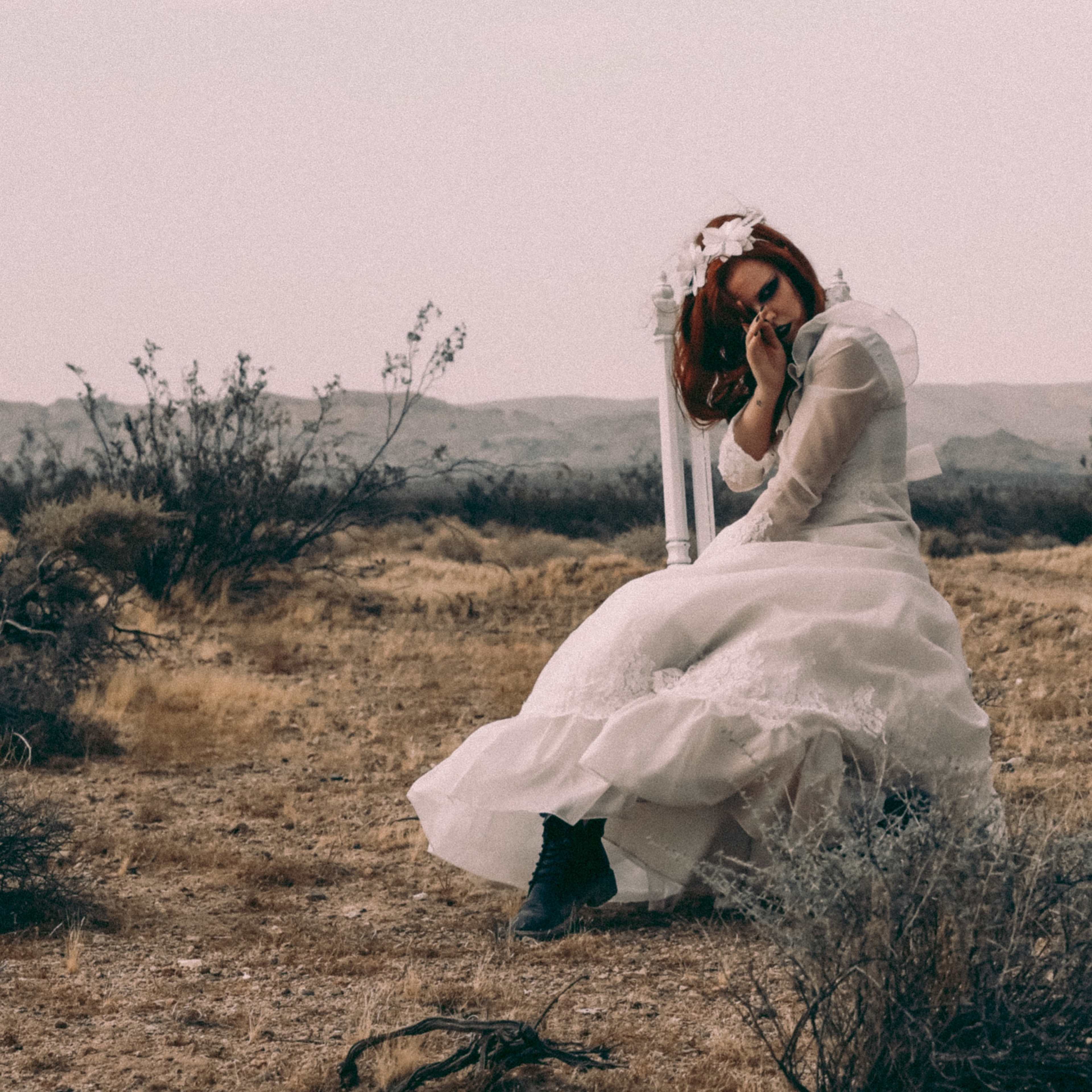 A woman in a wedding dress sits on the ground in a barren desert landscape, surrounded by sparse vegetation.