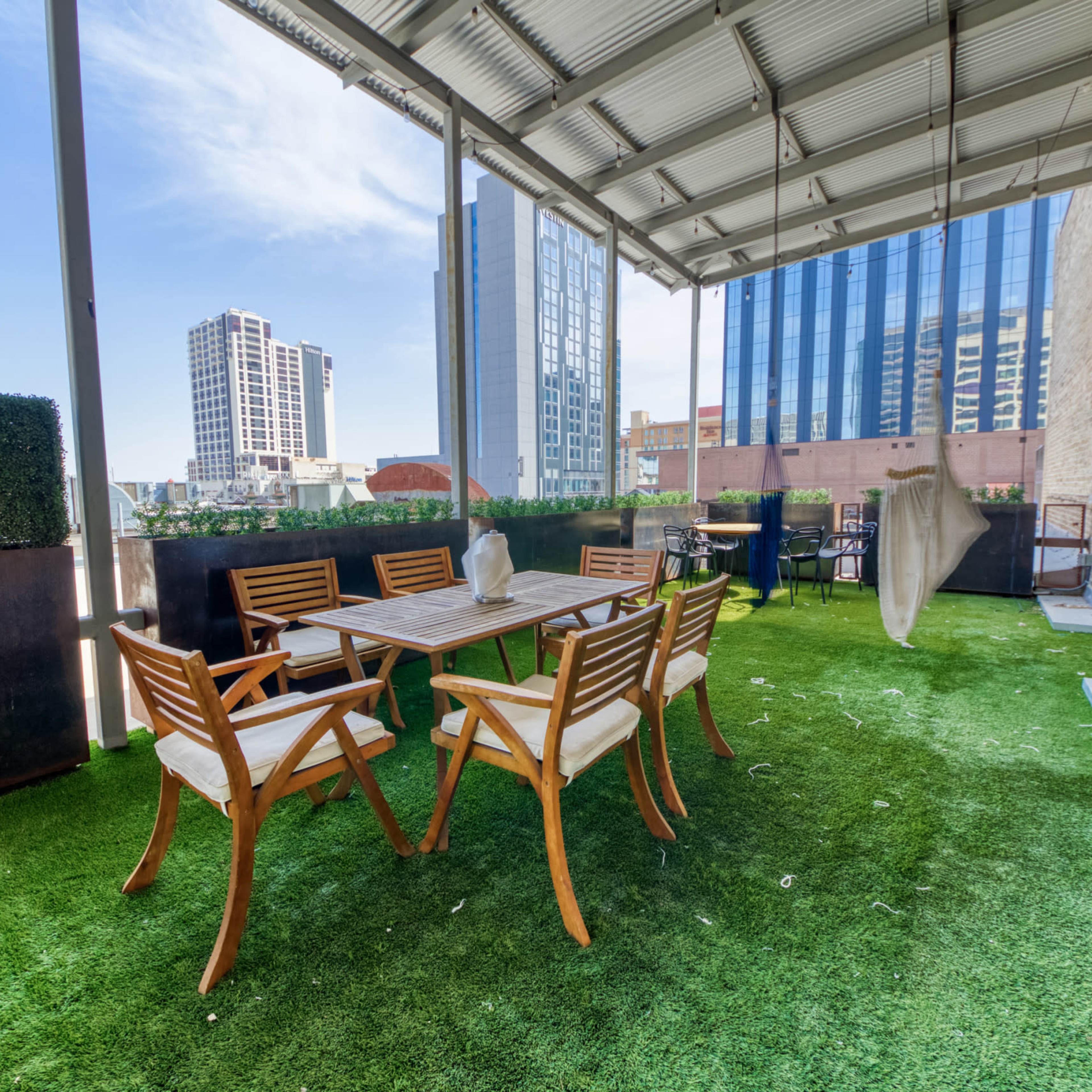 A rooftop terrace features a wooden dining table surrounded by chairs, with artificial grass and a backdrop of city skyscrapers.