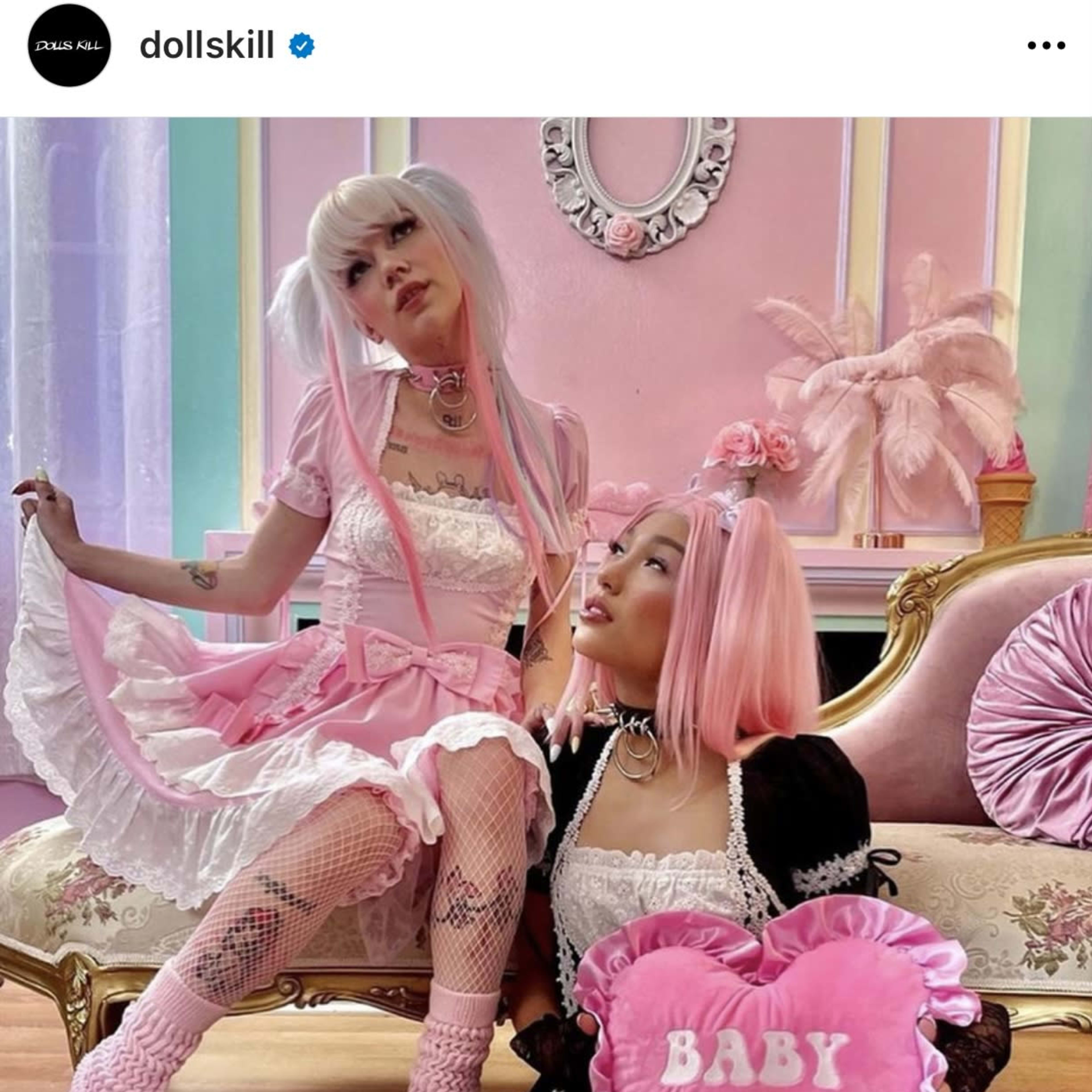 Two young women dressed in pastel-colored outfits and platform shoes pose on a vintage sofa in a brightly decorated room.
