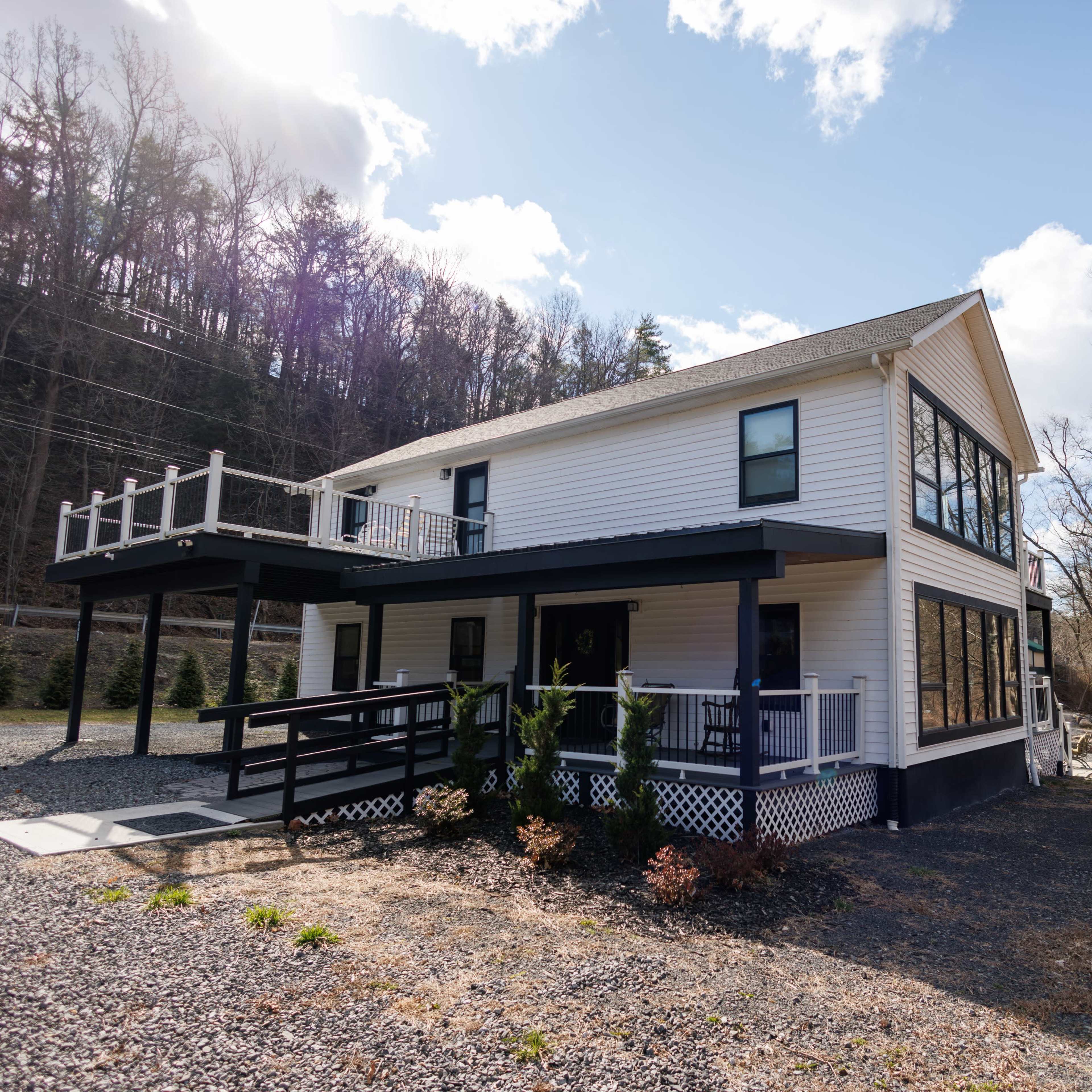 A modern two-story white house with large windows and a balcony, situated near a gravel driveway alongside a river.