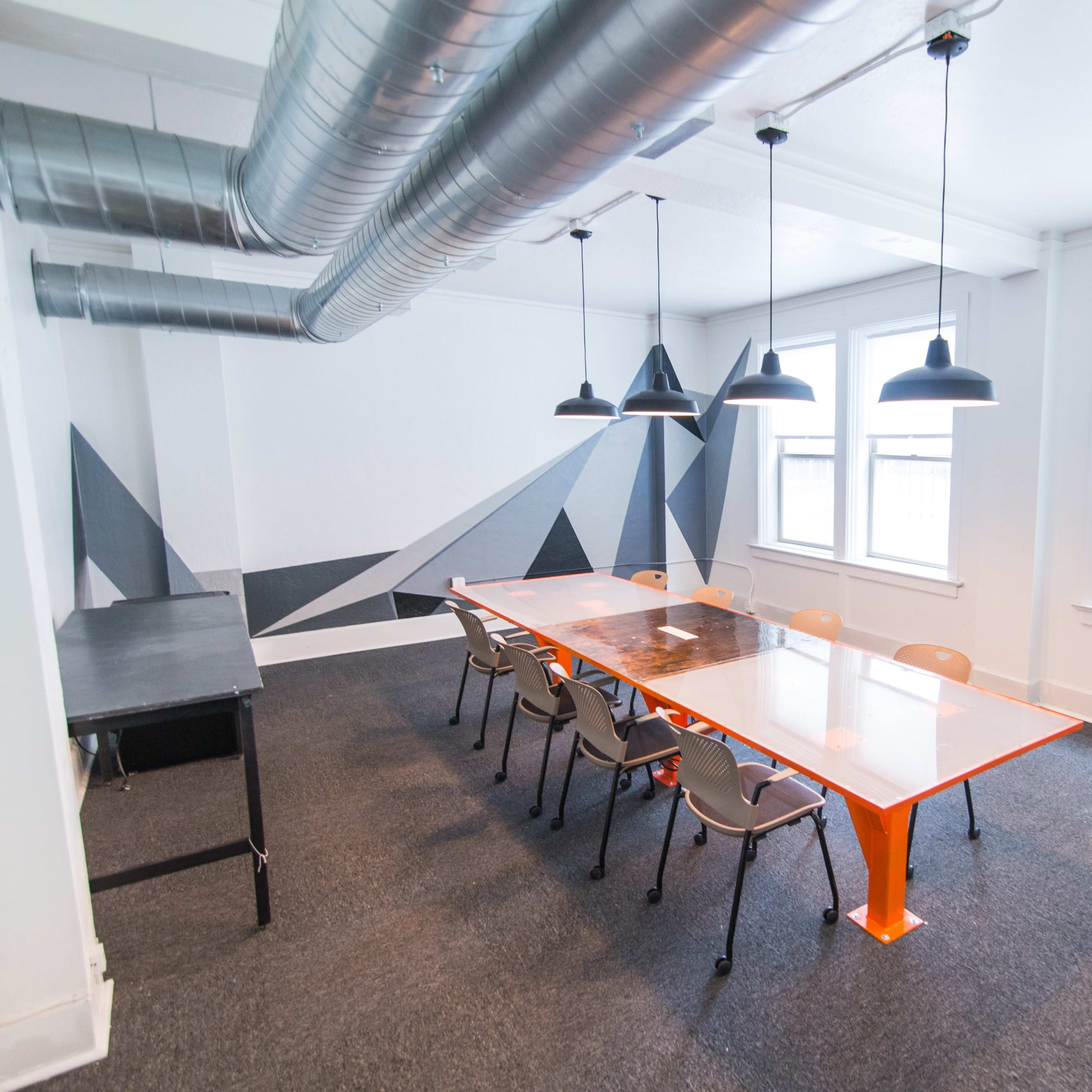 A spacious conference room features a long glass table with orange legs, surrounded by eight gray chairs, and is illuminated by pendant lights with a geometric mural on the wall.