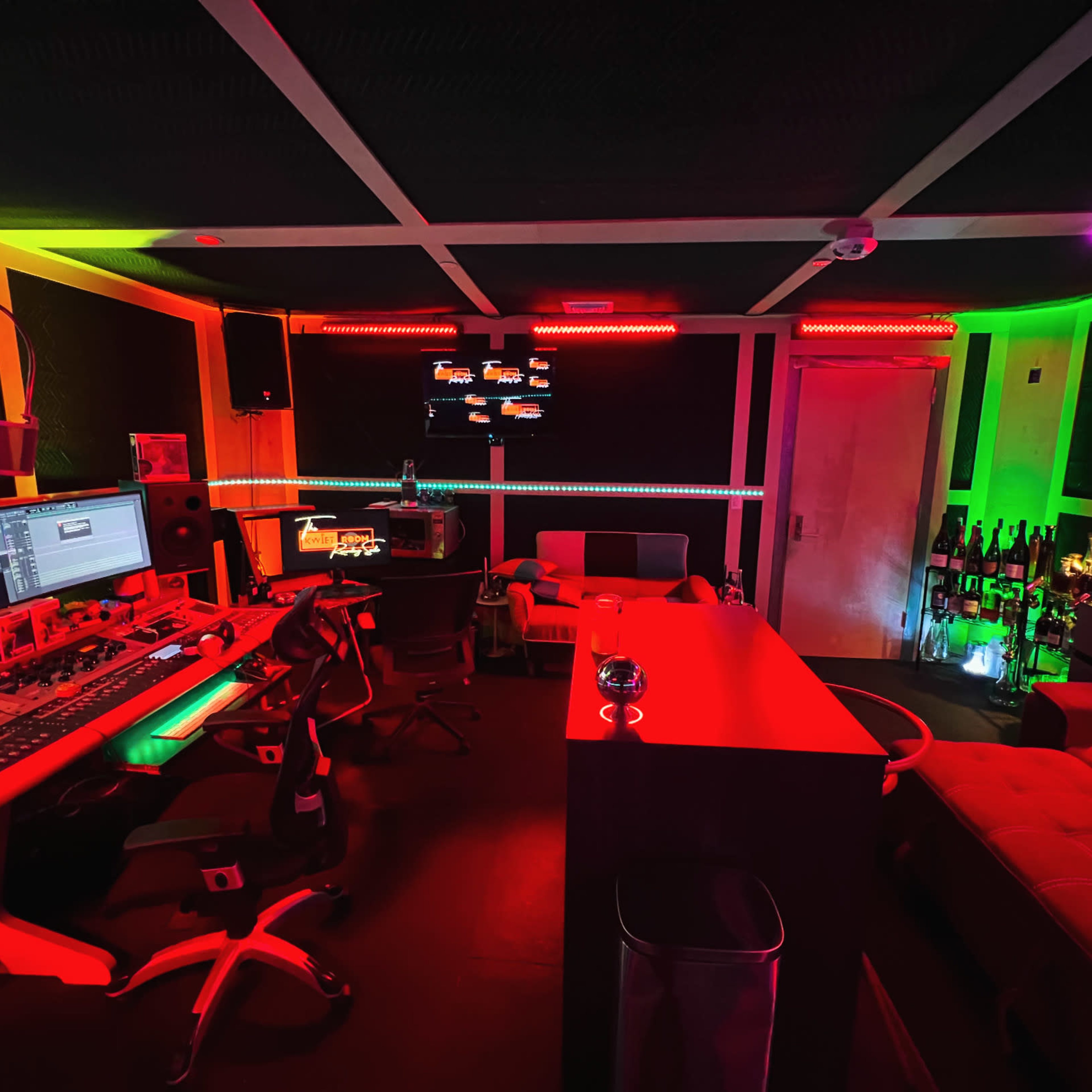 A music studio with colorful LED lighting, equipped with a mixing console, computer monitors, and a lounge area featuring a bar and seating.