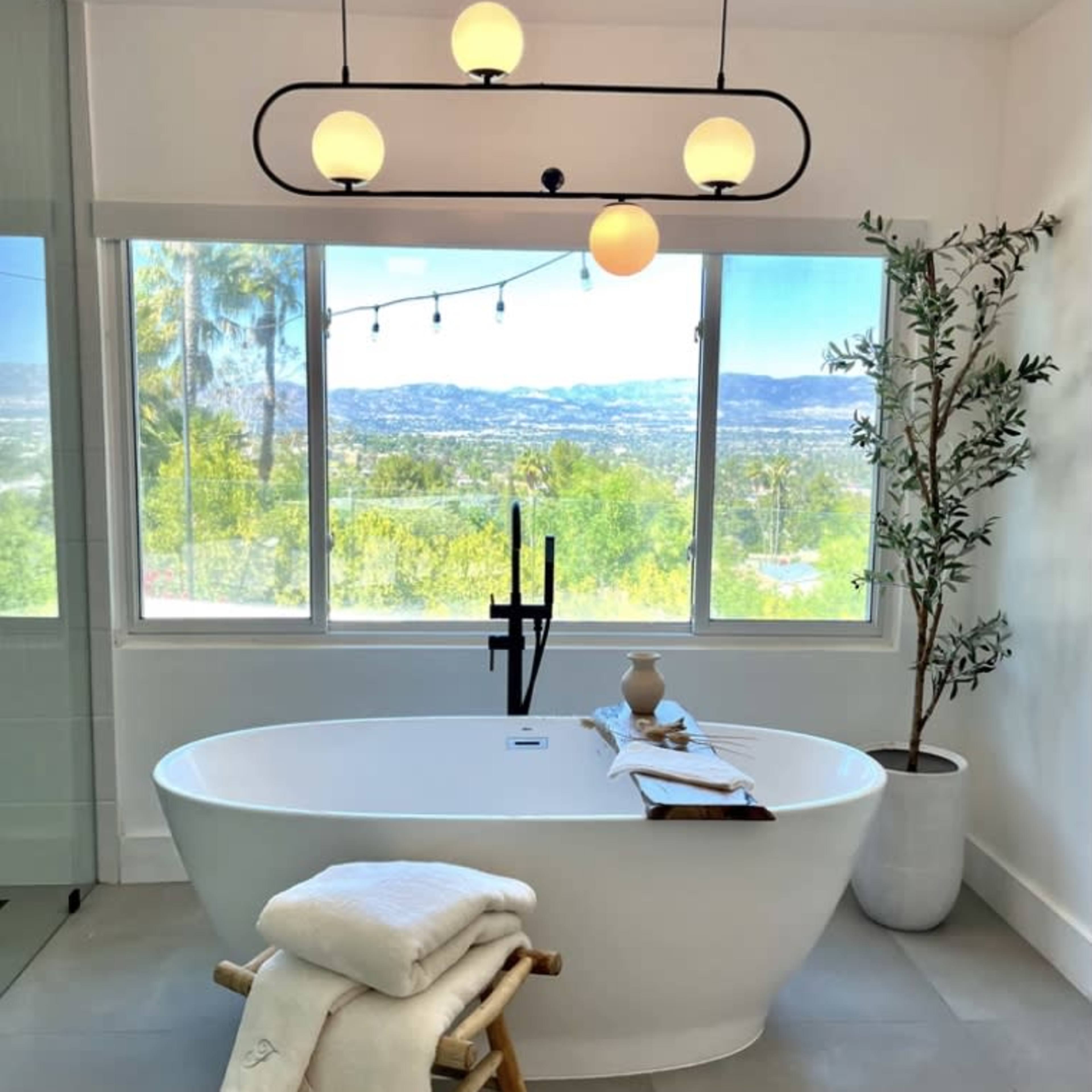 A modern freestanding bathtub is positioned in front of a large window offering a view of the outdoors, with a small table beside it holding a towel and decorative items.