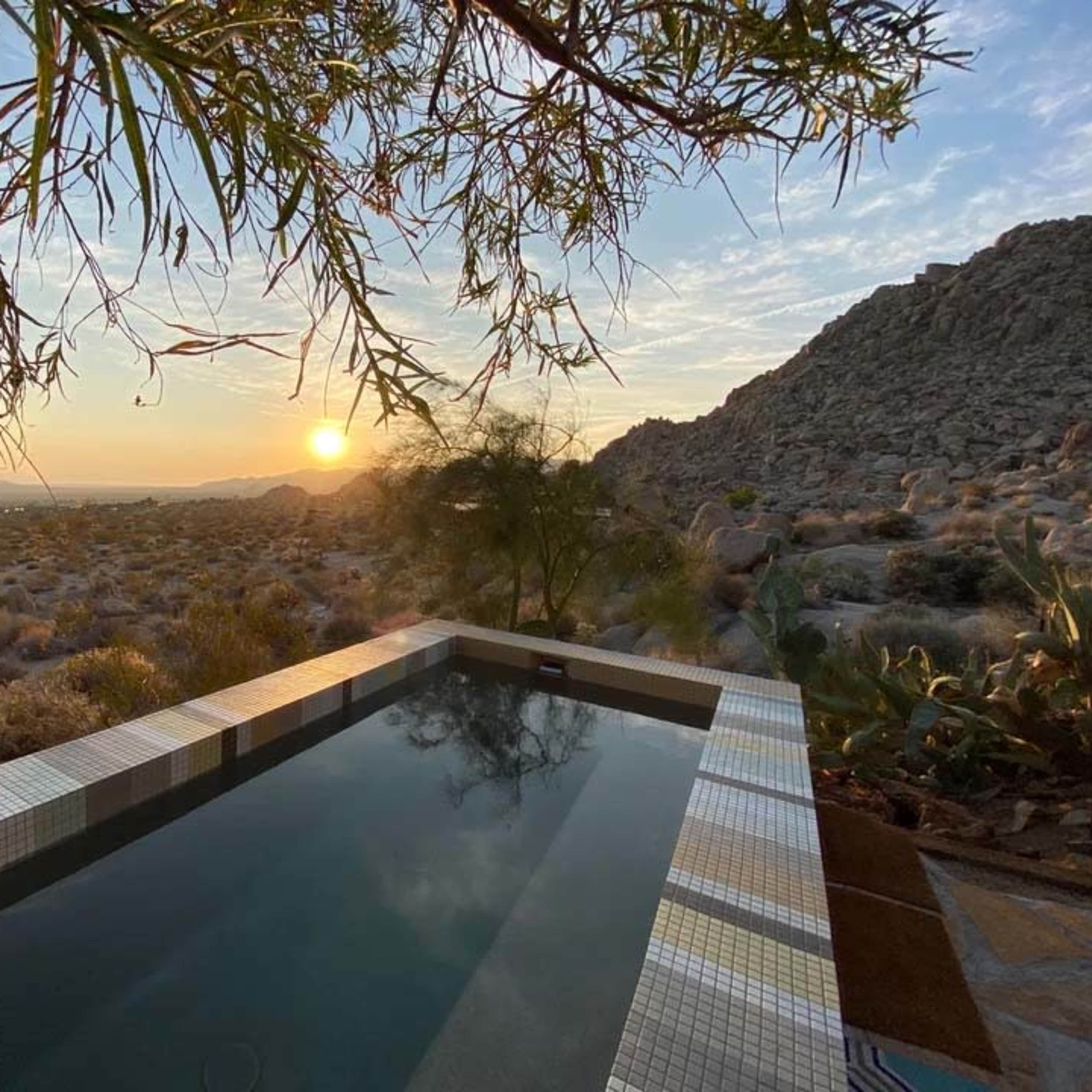 A rectangular pool with tiled edges reflects the sunset over a desert landscape framed by trees and rocky hills.
