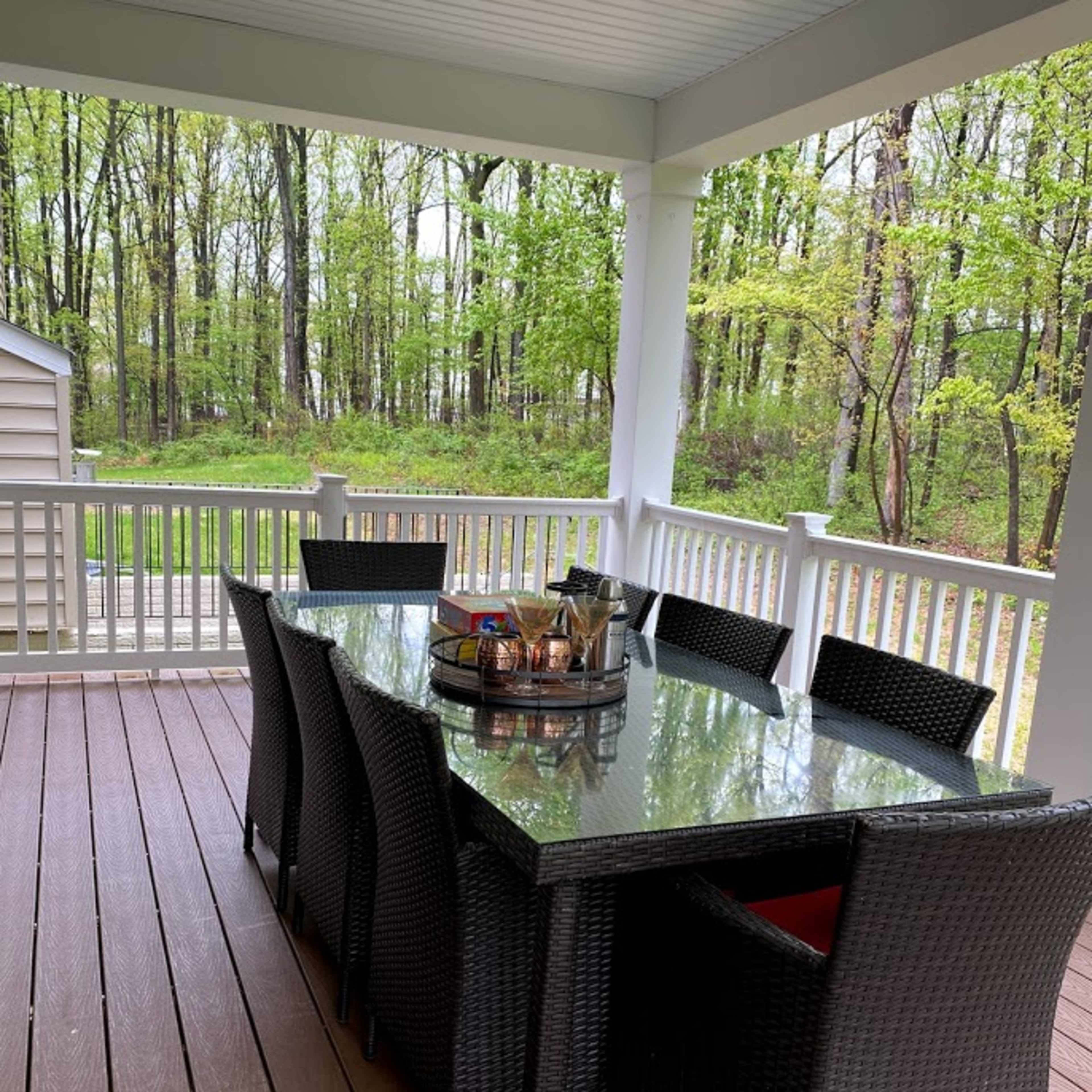 A rectangular dining table with eight chairs sits on a covered porch surrounded by trees.