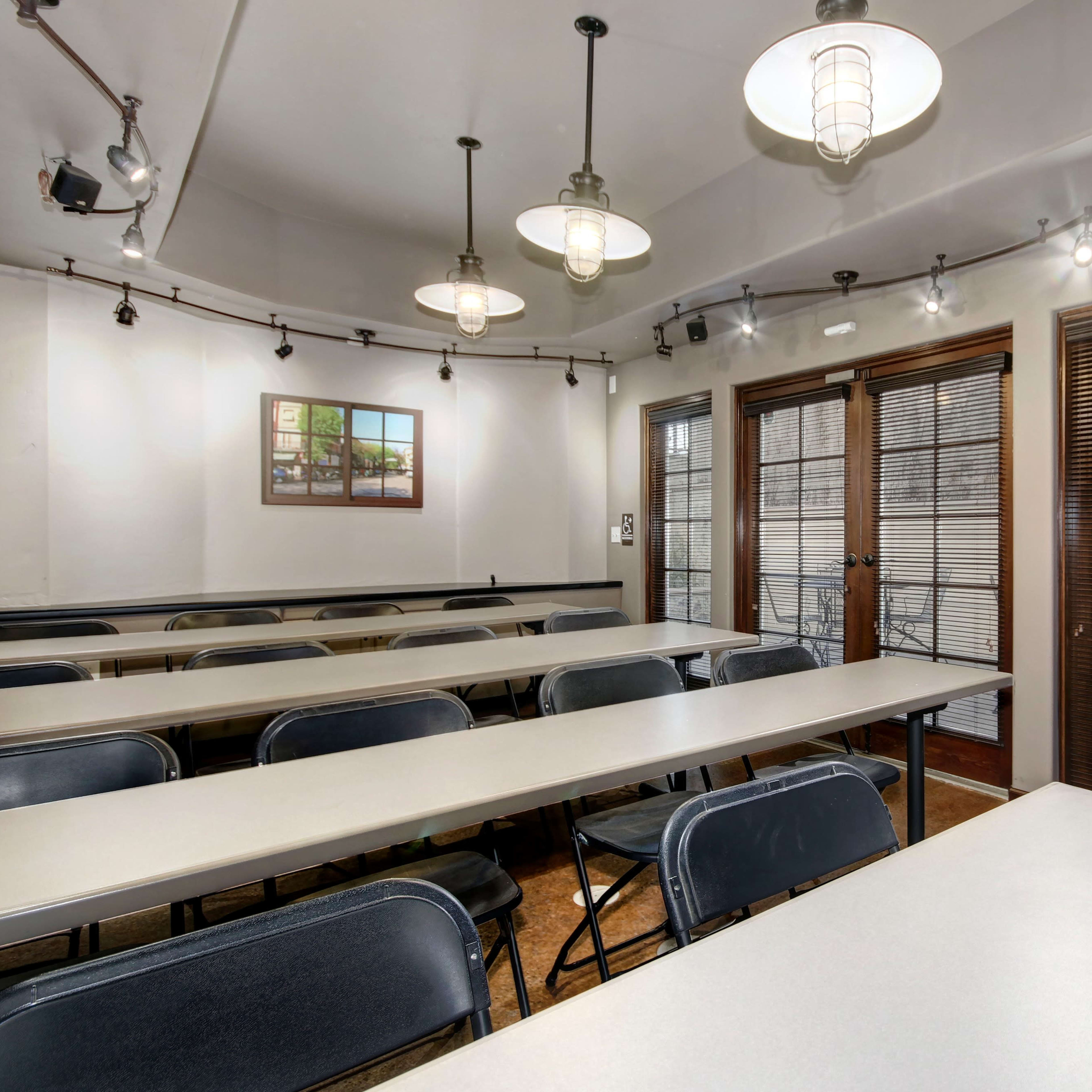A classroom setting with several rows of tables and chairs, illuminated by overhead lights, and large windows featuring blinds.