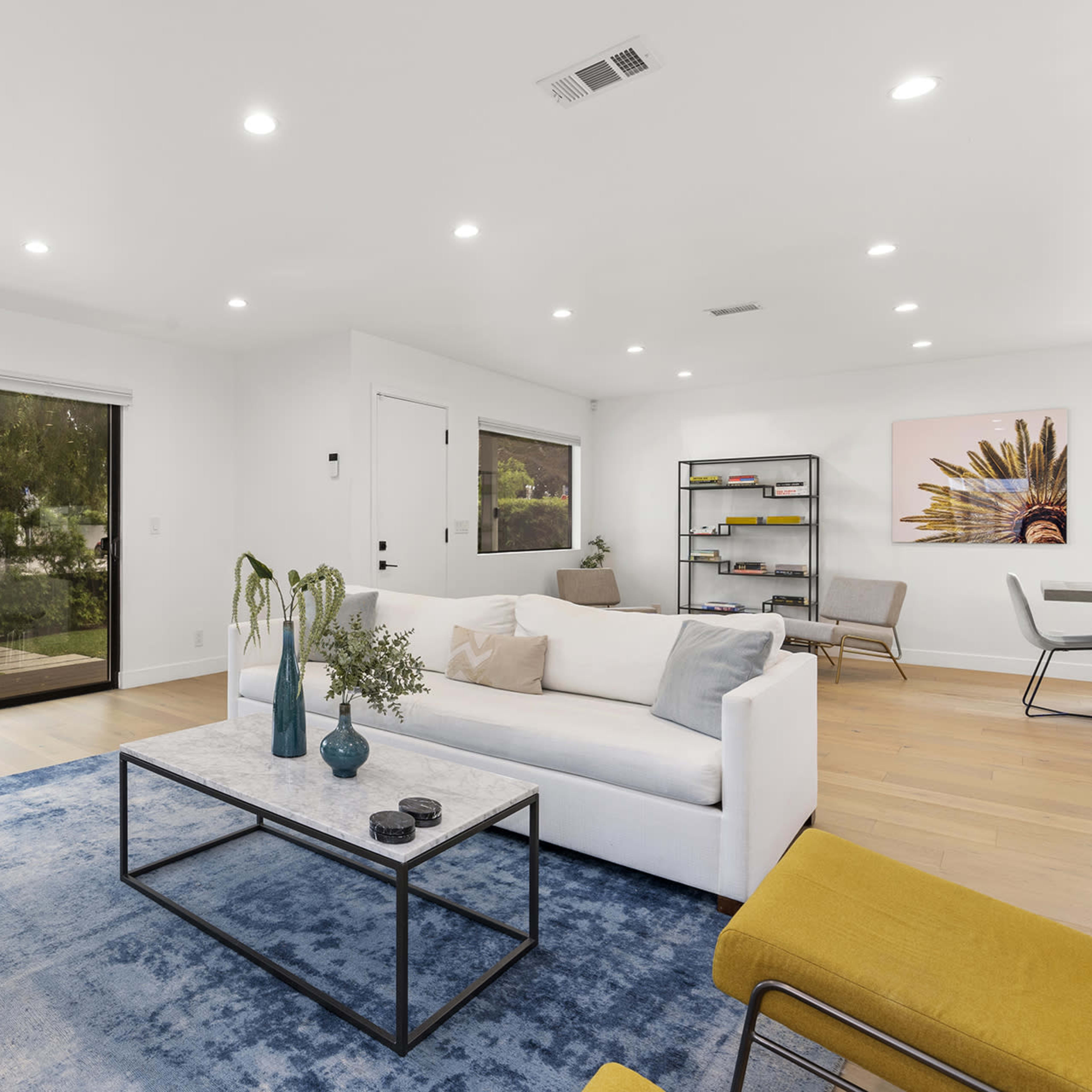 A modern living room features a white sofa, a glass coffee table, and a brown armchair, with large windows overlooking a garden area.