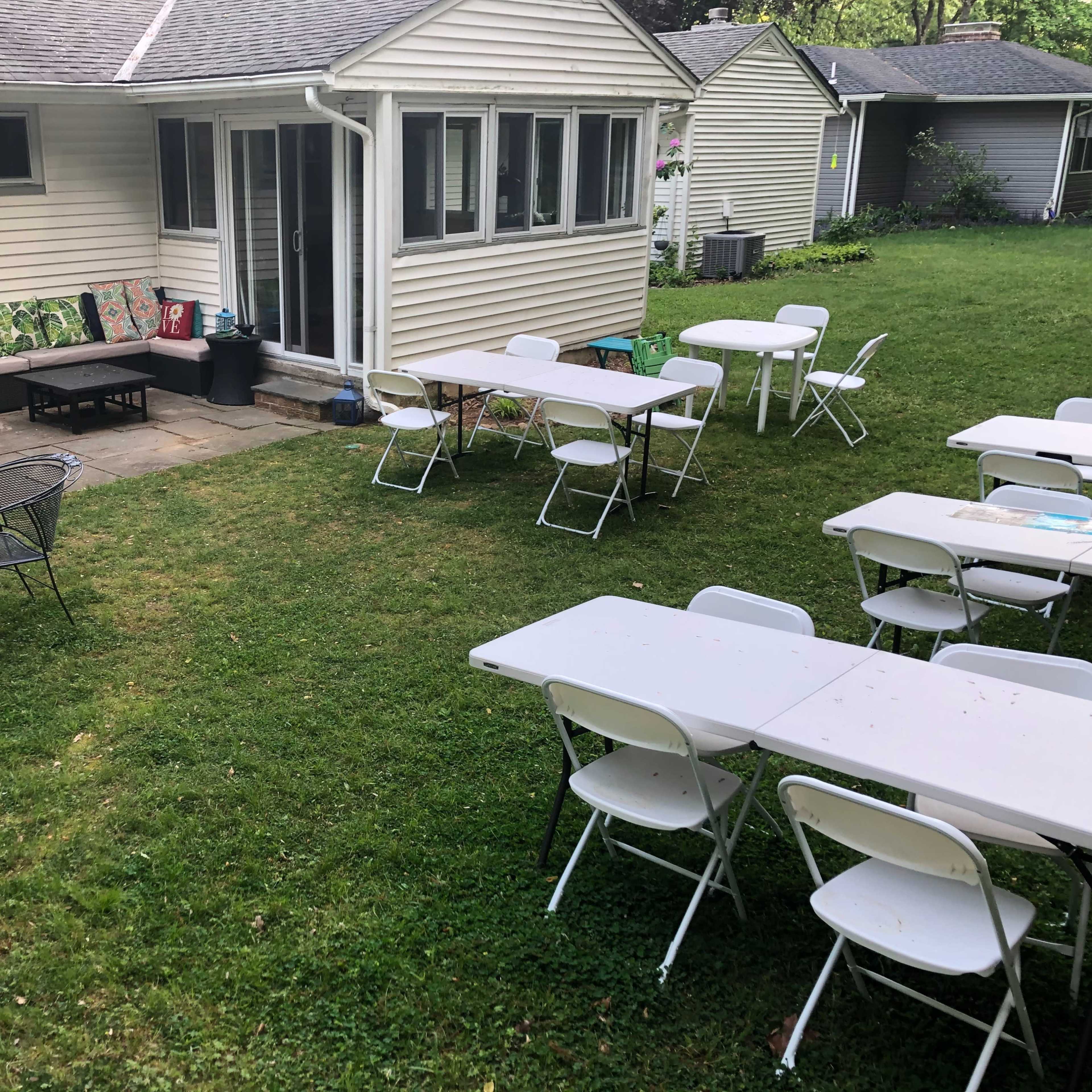 A grassy backyard features several empty folding tables and chairs arranged for an outdoor gathering, with a house visible in the background.