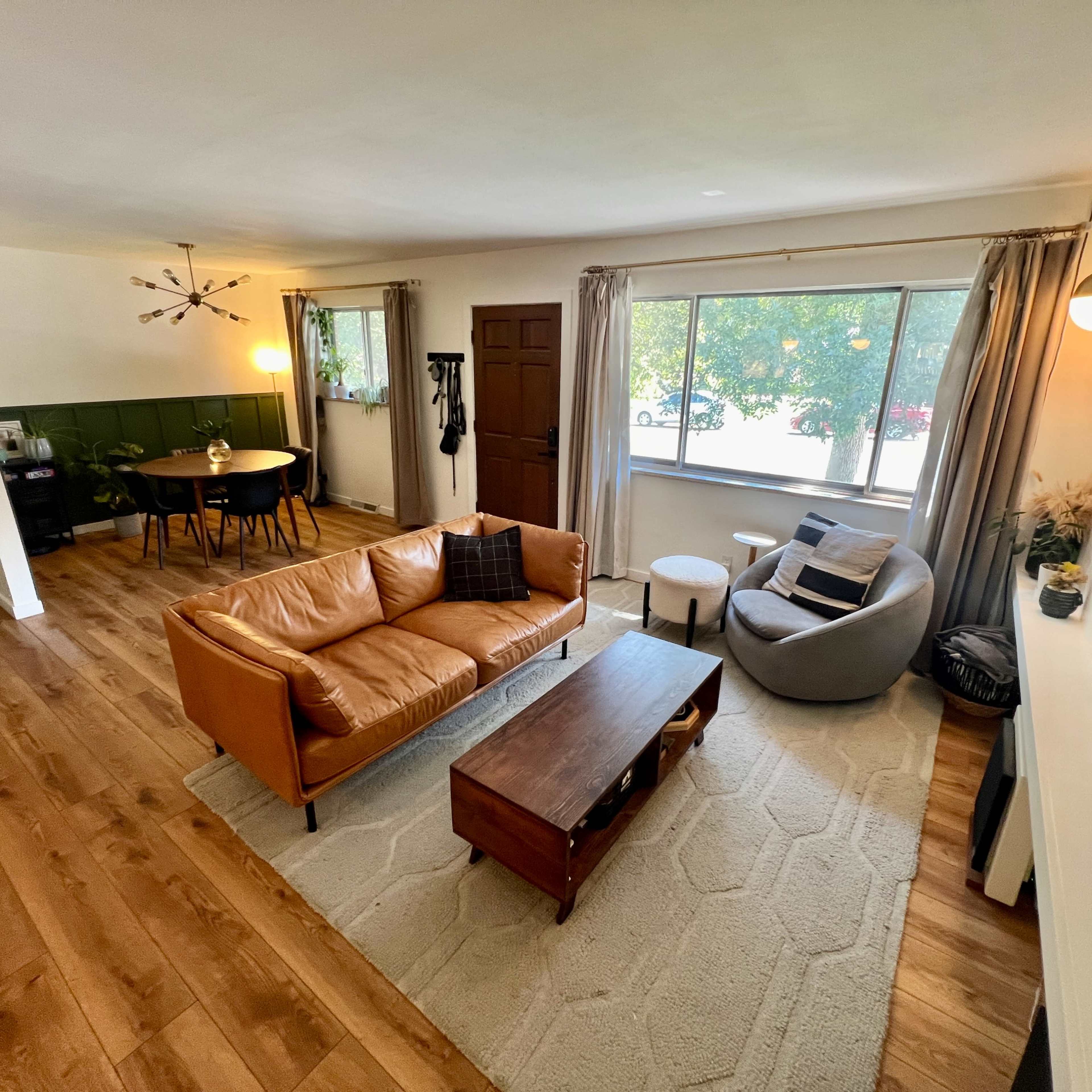 A living room with a brown leather sofa, a circular chair, a wooden coffee table, and a large window letting in natural light.