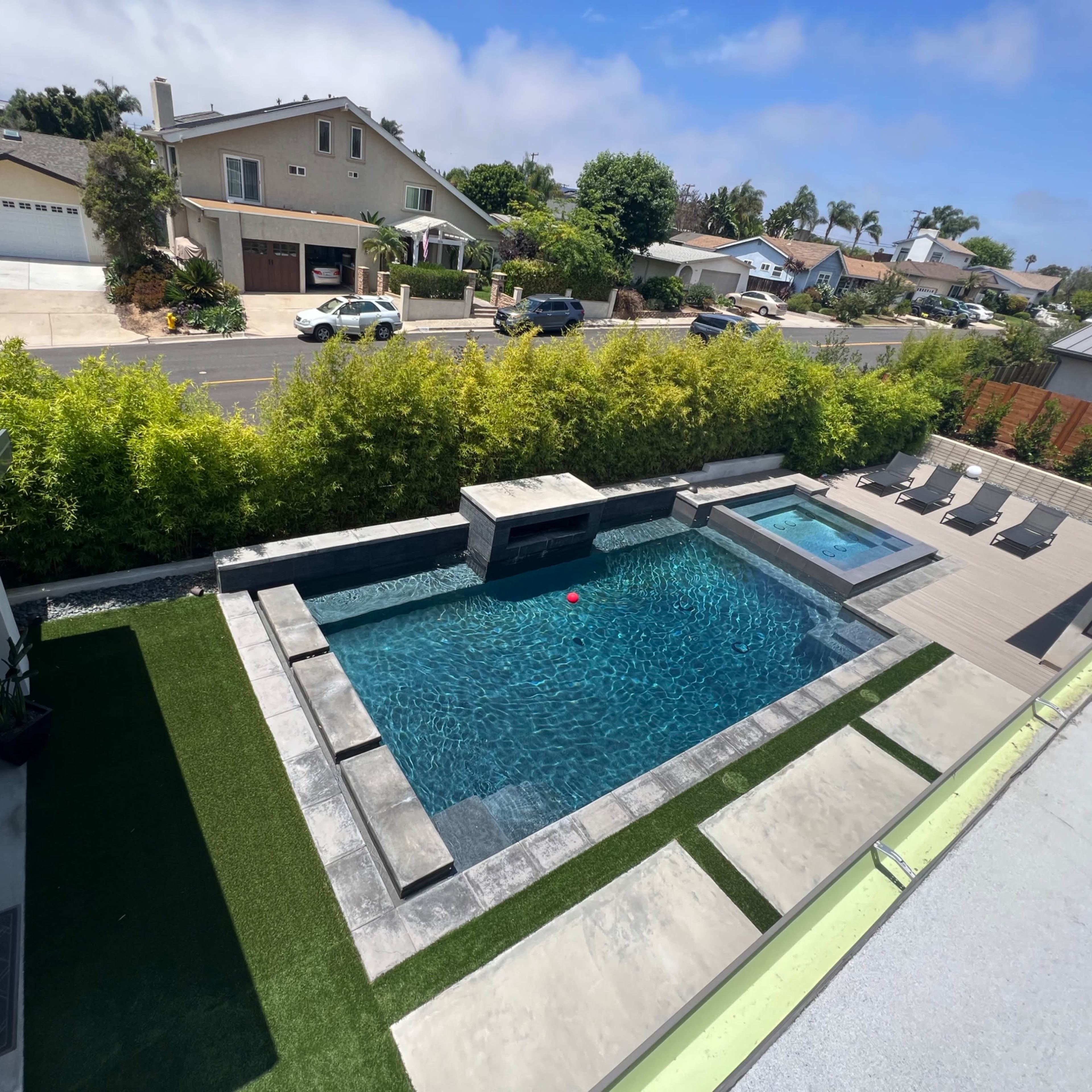 A landscaped backyard featuring a rectangular swimming pool, a spa, and several lounge chairs, with a neighborhood visible in the background.