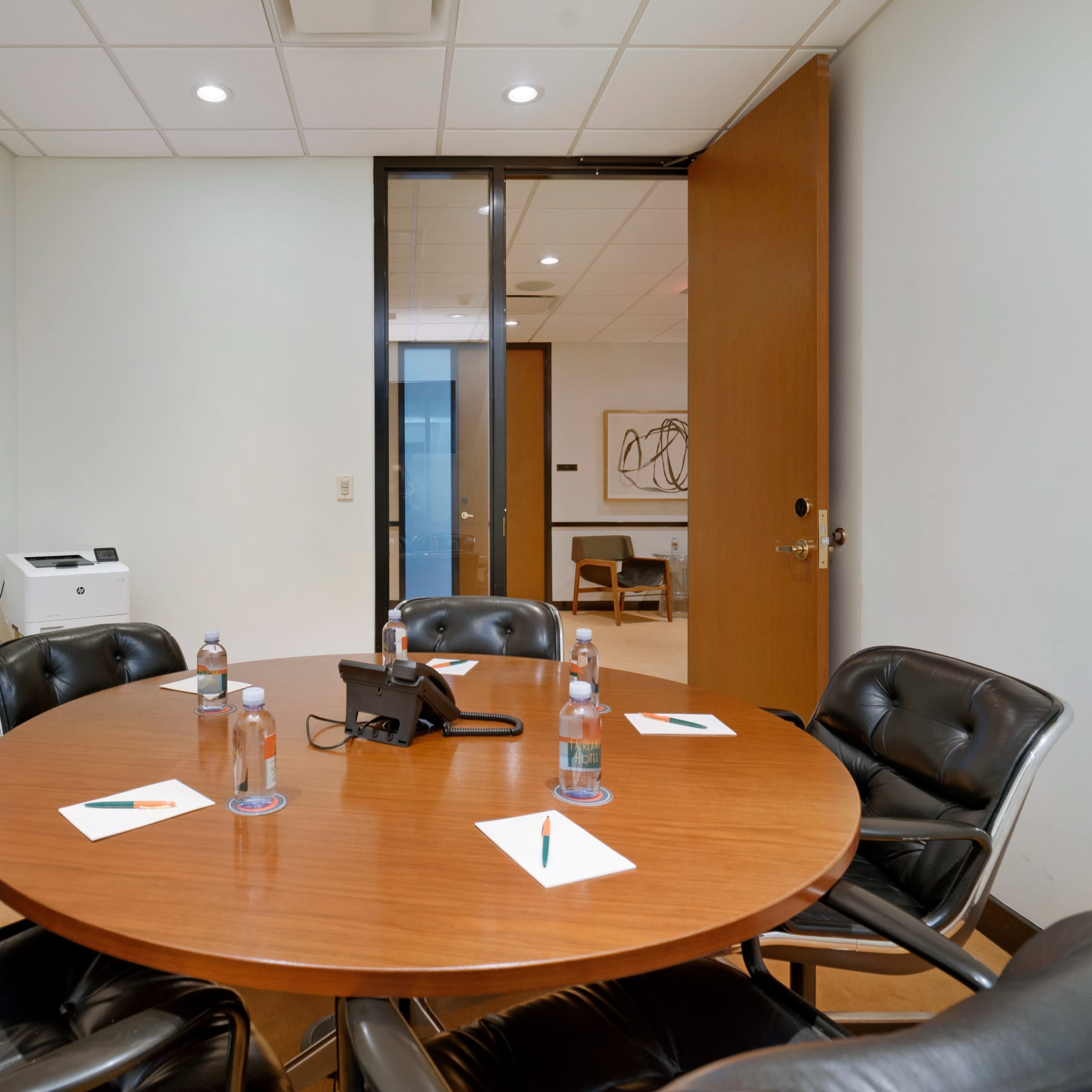 A round wooden table with black chairs and a phone is set up in a conference room, featuring water bottles and notepads at each seat.