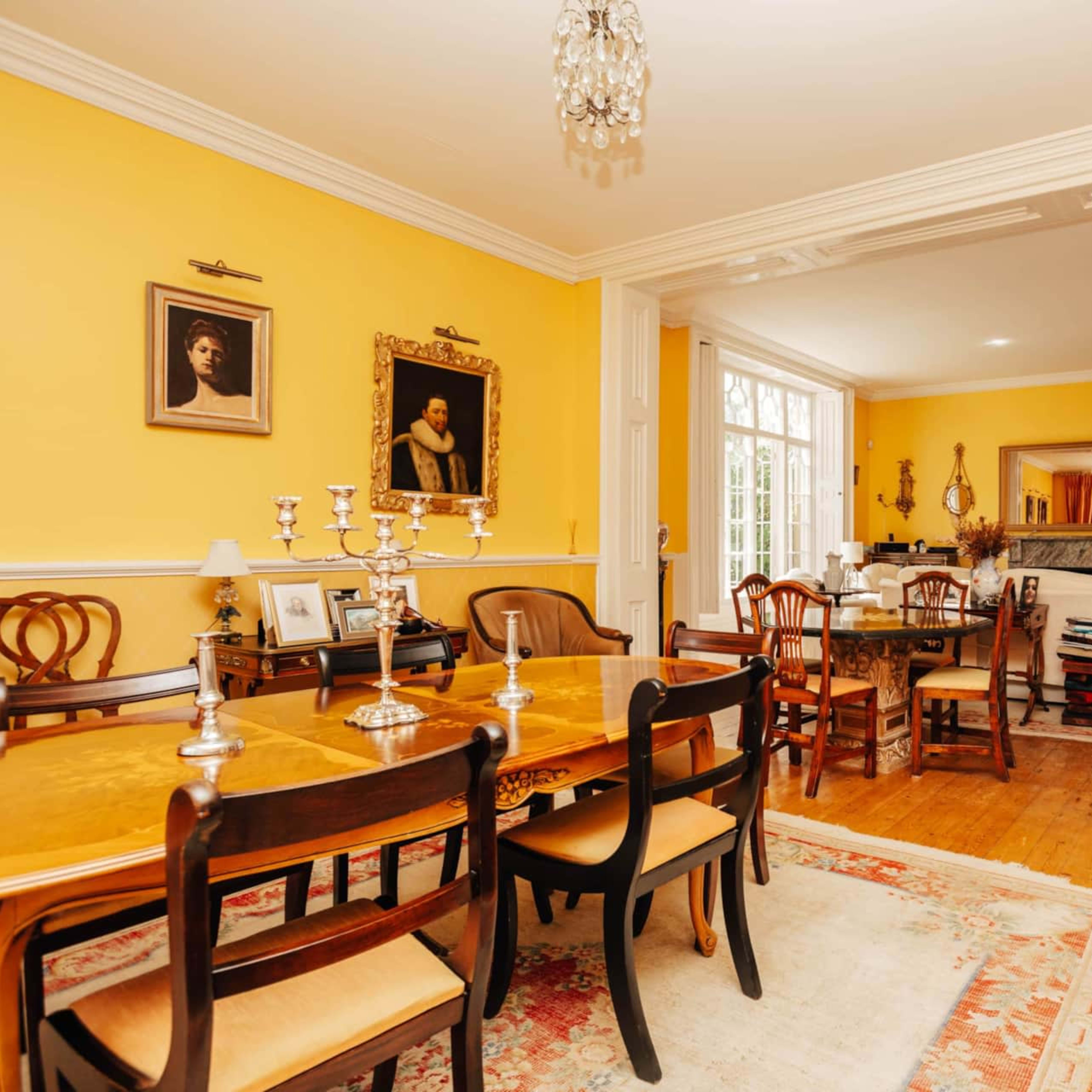 A brightly lit dining room featuring a wooden dining table surrounded by chairs, with yellow walls adorned with framed portraits and a chandelier overhead.