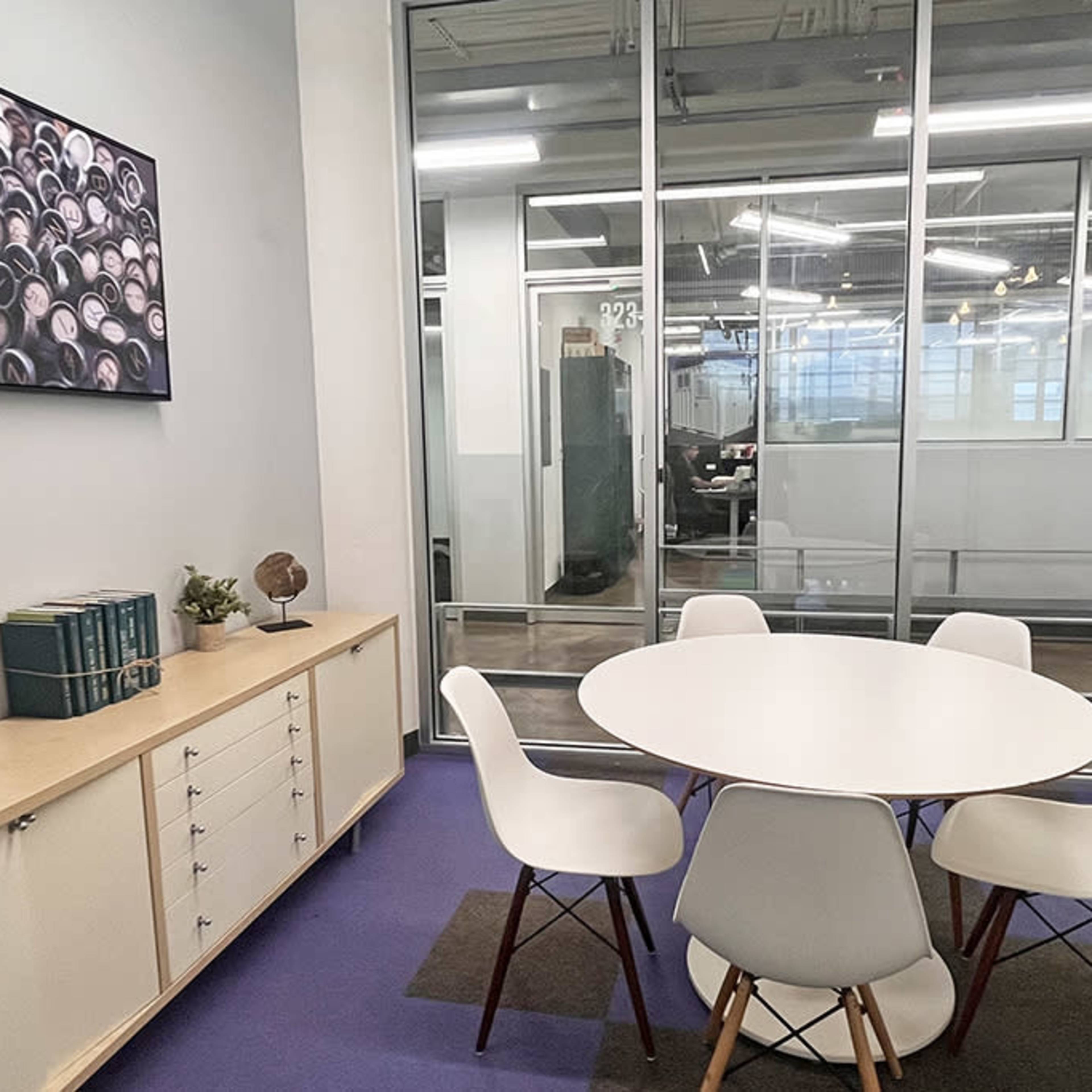 A small meeting room with a round table surrounded by four white chairs, a cabinet with books and a globe, and glass walls providing a view of an office space beyond.