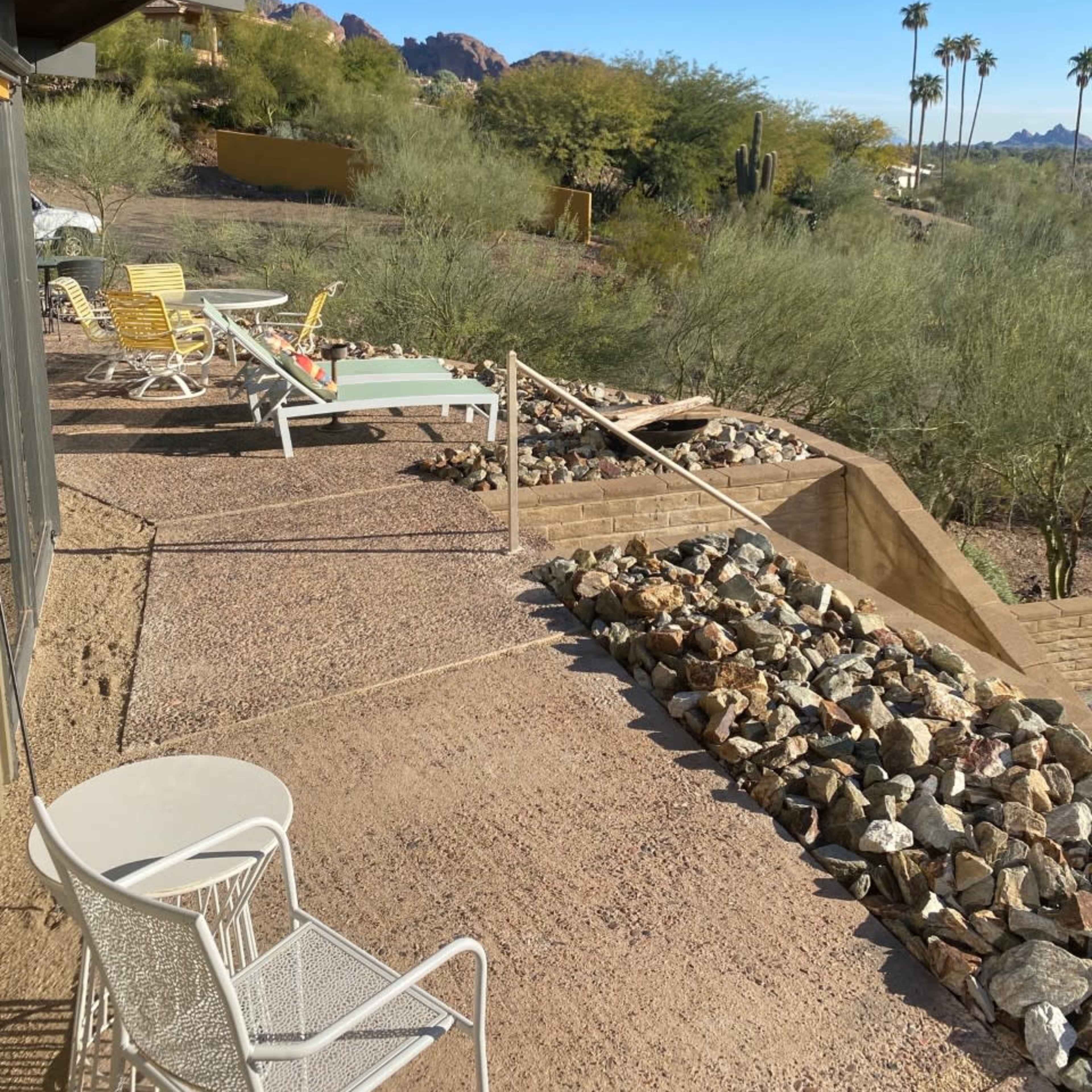 A patio area features lounge chairs and a small table, with a rocky pathway leading to a landscaped yard and desert vegetation in the background.