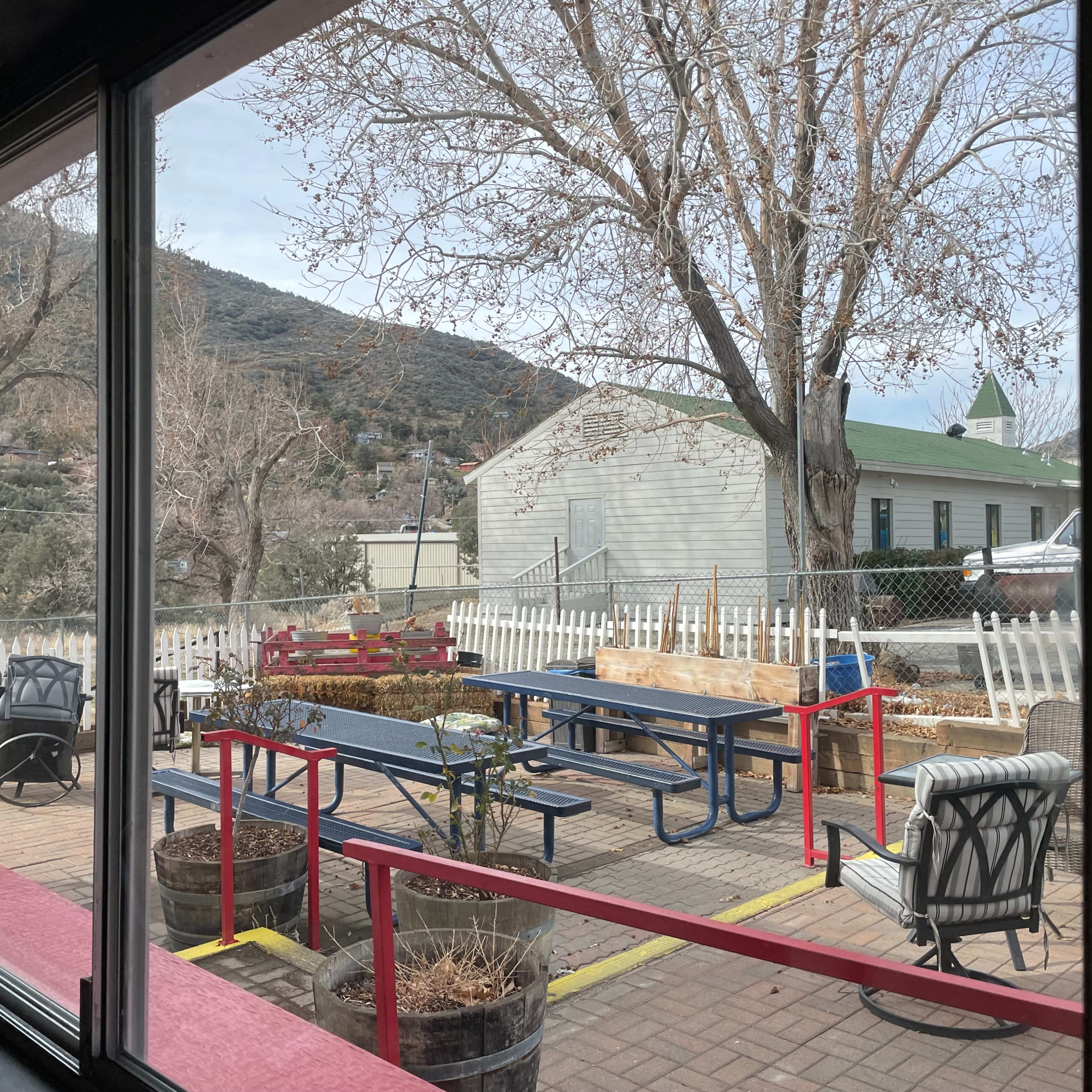 An outdoor seating area with picnic tables and chairs, viewed through a window, against a backdrop of bare trees and a building with a green roof.