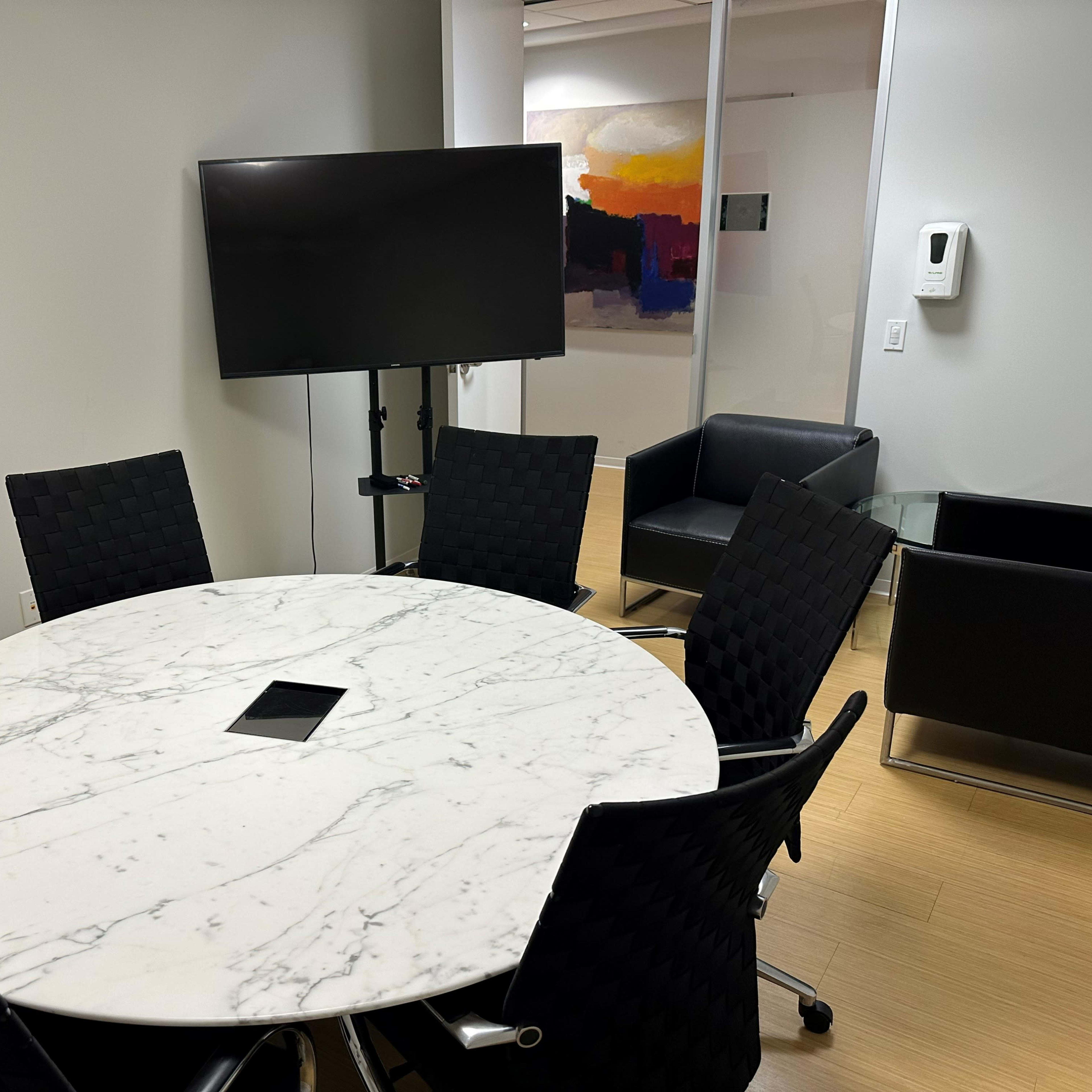 A round marble conference table with black chairs is set in a room featuring a television mounted on a stand and a black leather chair in the corner.