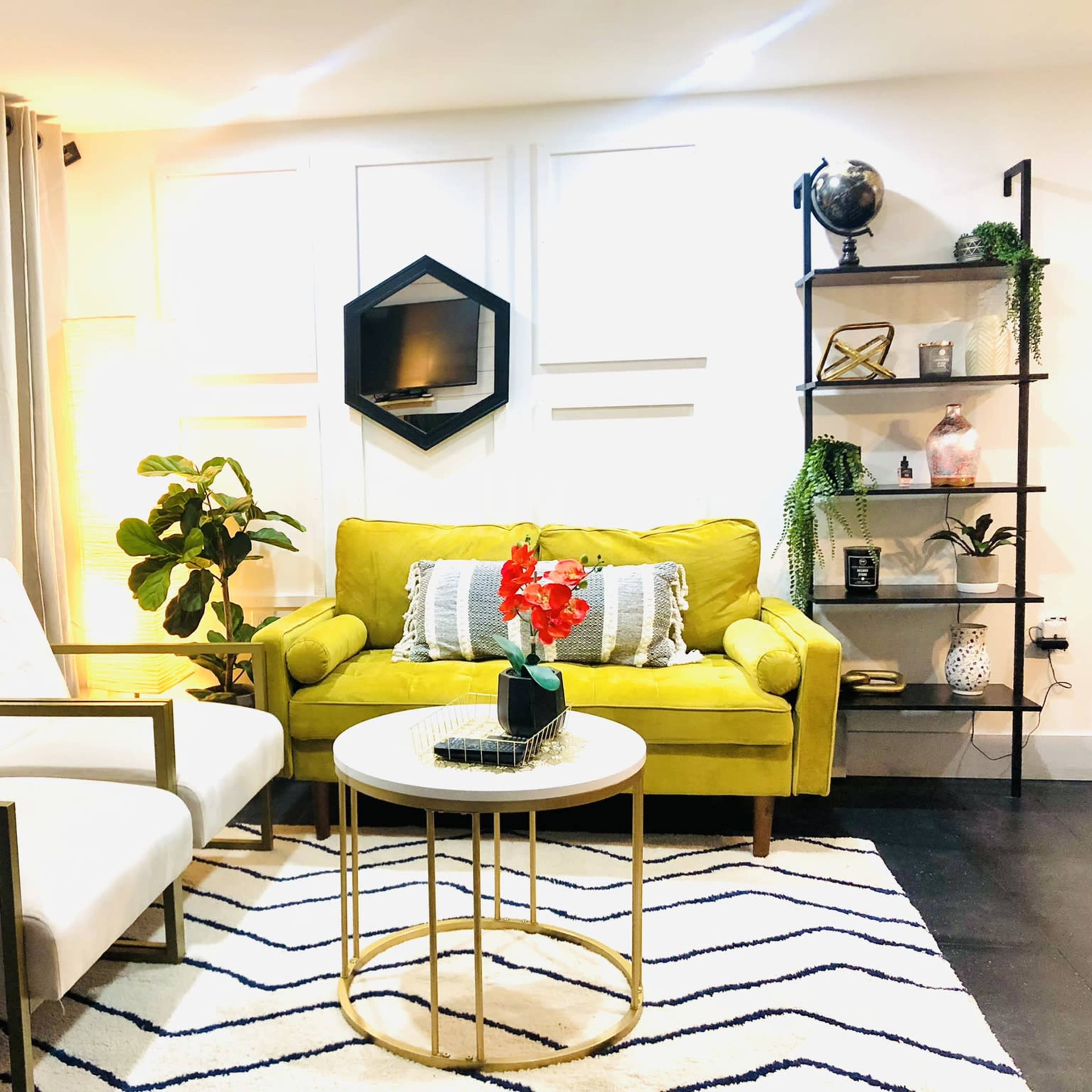 A cozy living room featuring a green sofa, two white chairs, a round coffee table, and decorative shelves with plants and accessories.