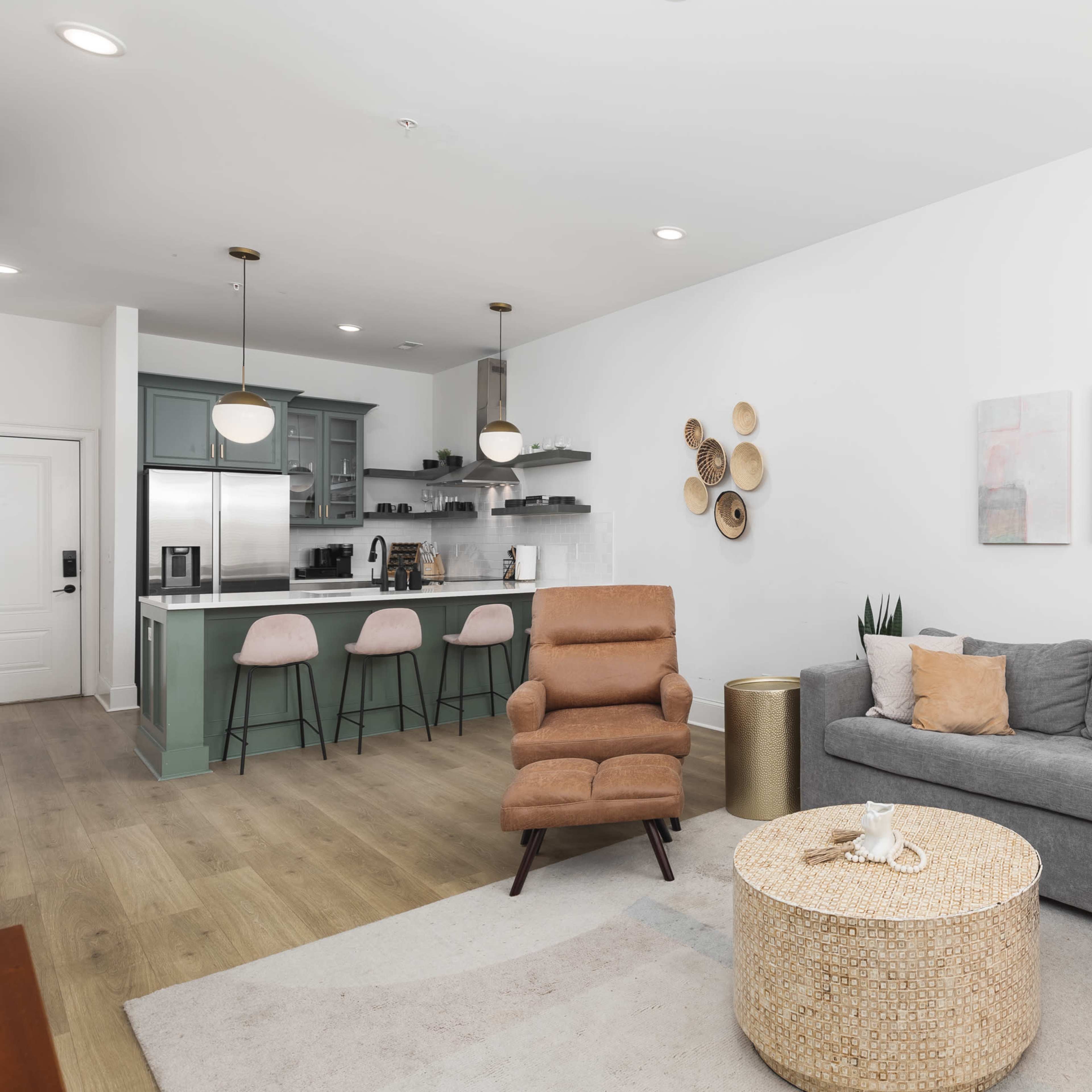 The living area features a gray sofa, a brown recliner, a round woven coffee table, and a kitchen with bar stools in the background.