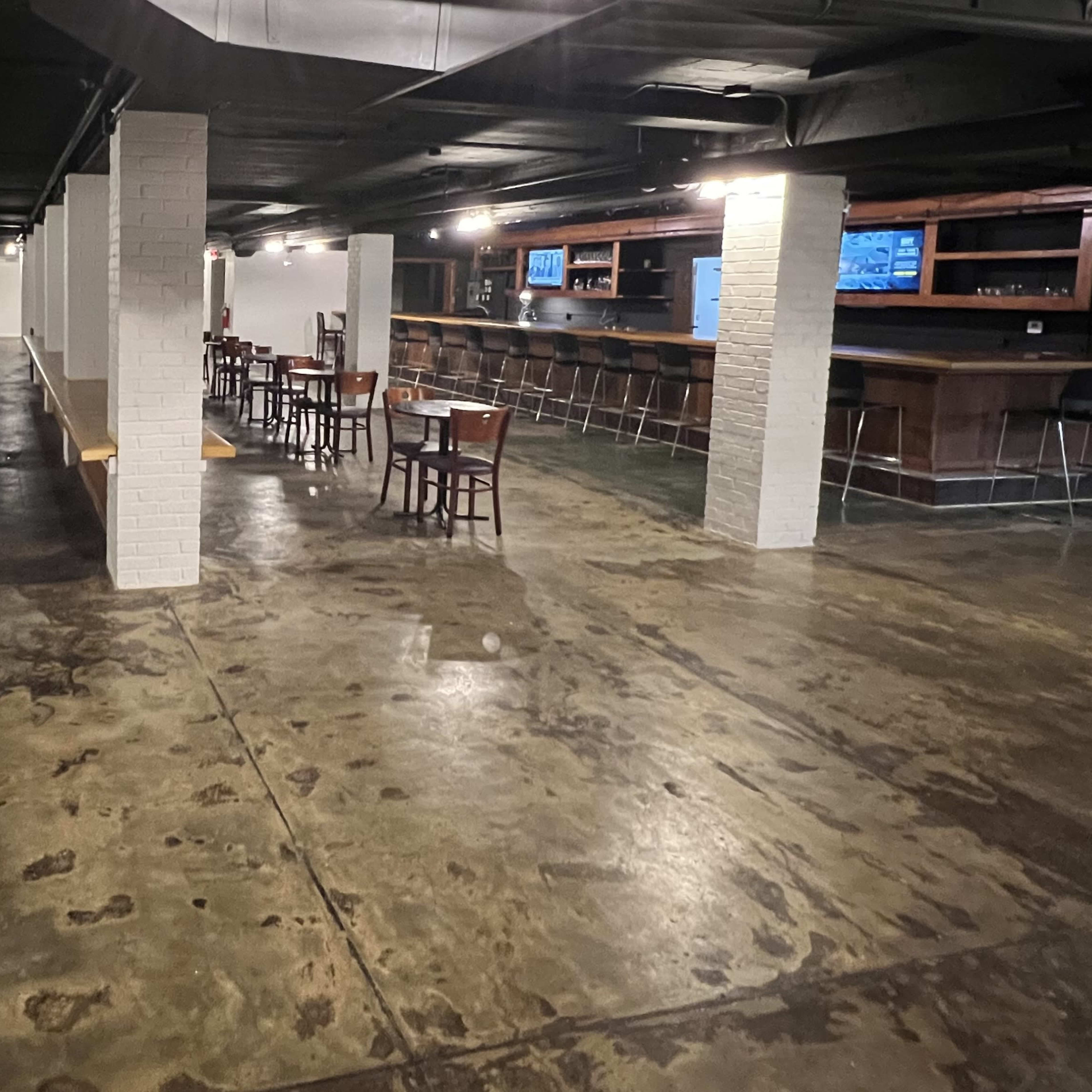 A spacious, dimly lit bar area features scattered wooden tables and a long counter against a concrete floor.