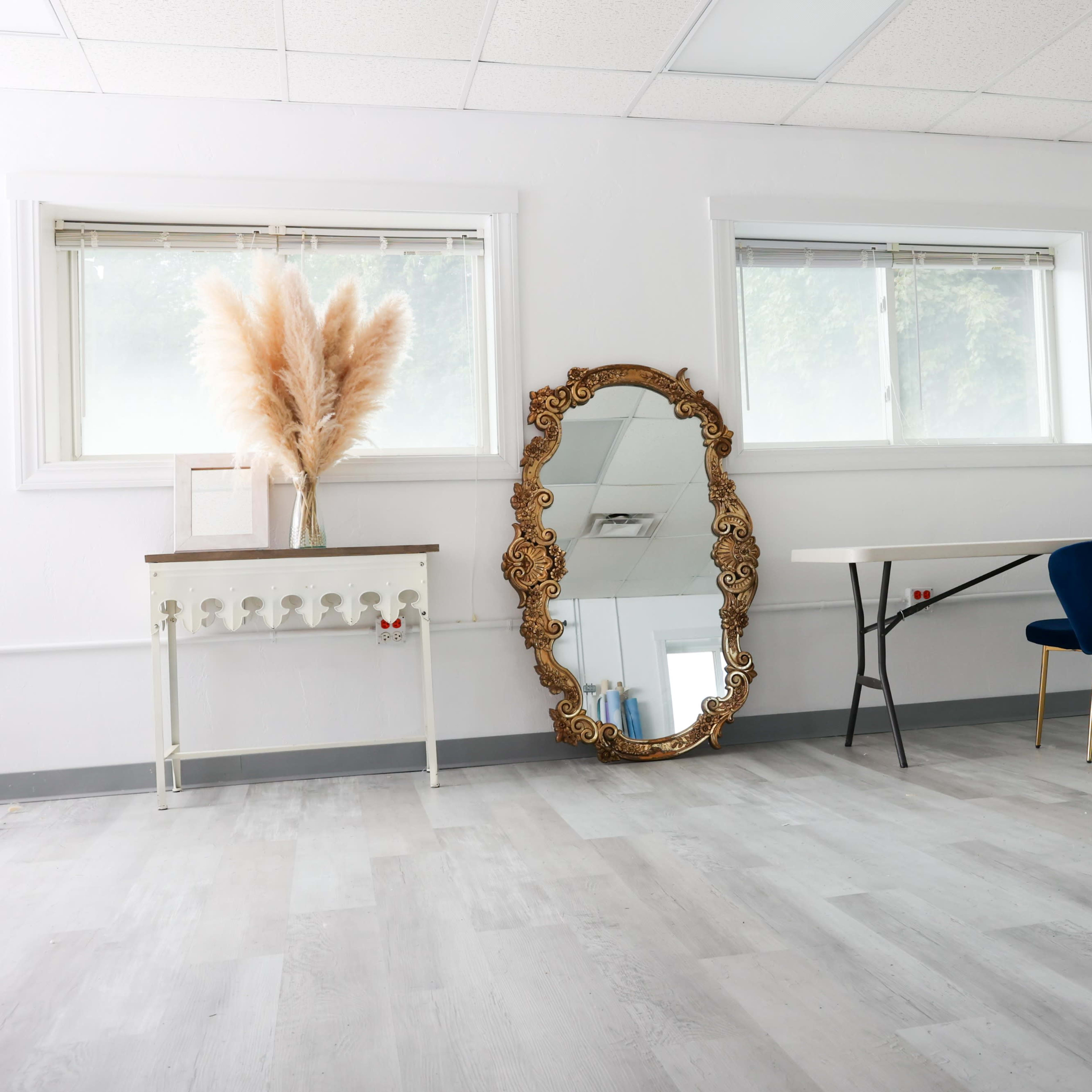 A bright, minimalist room featuring a large ornate mirror, a table with a vase of pampas grass, a blue chair, and a light reflector stand.