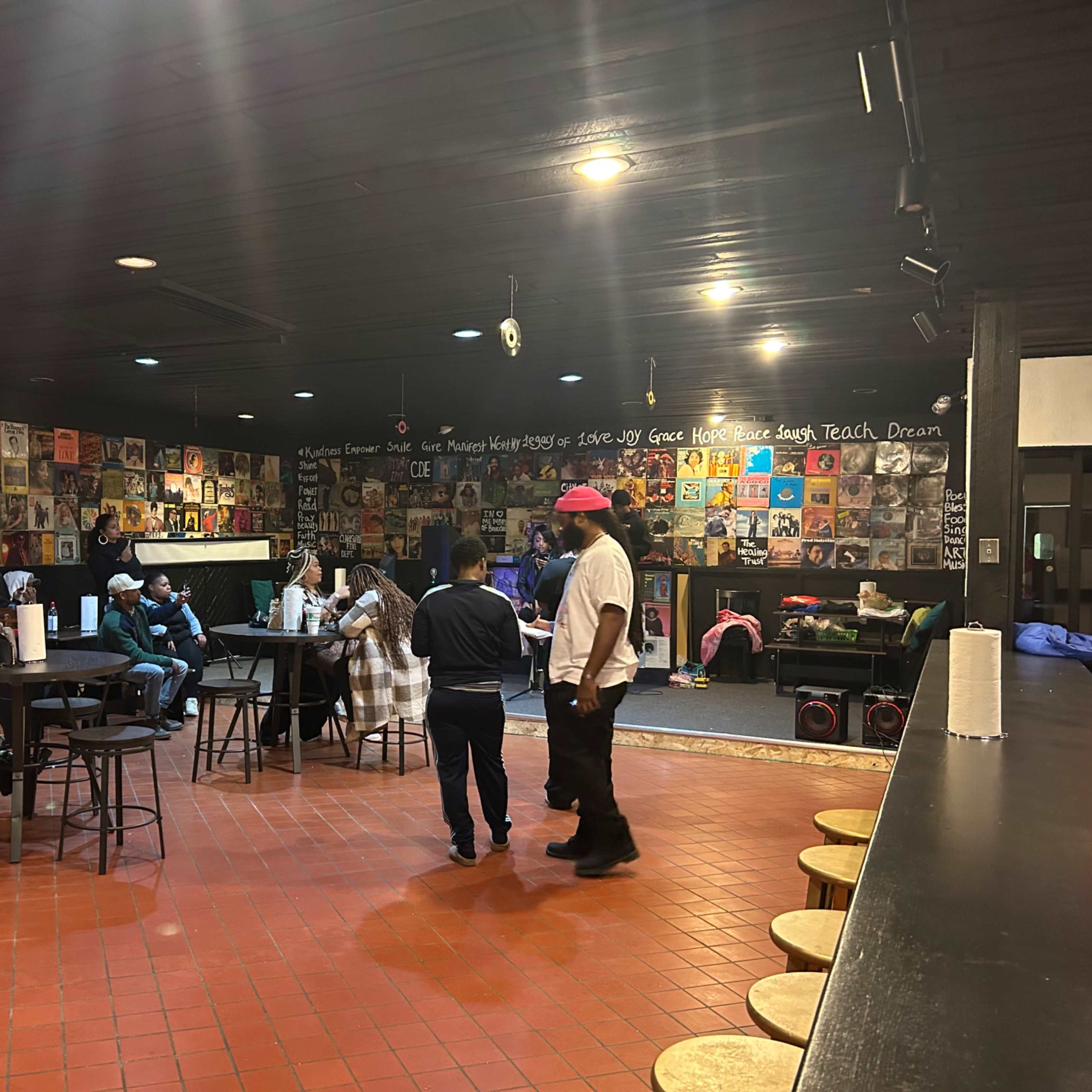 A dimly lit bar or lounge area with patrons seated at small tables, surrounded by walls decorated with vinyl records and large murals.