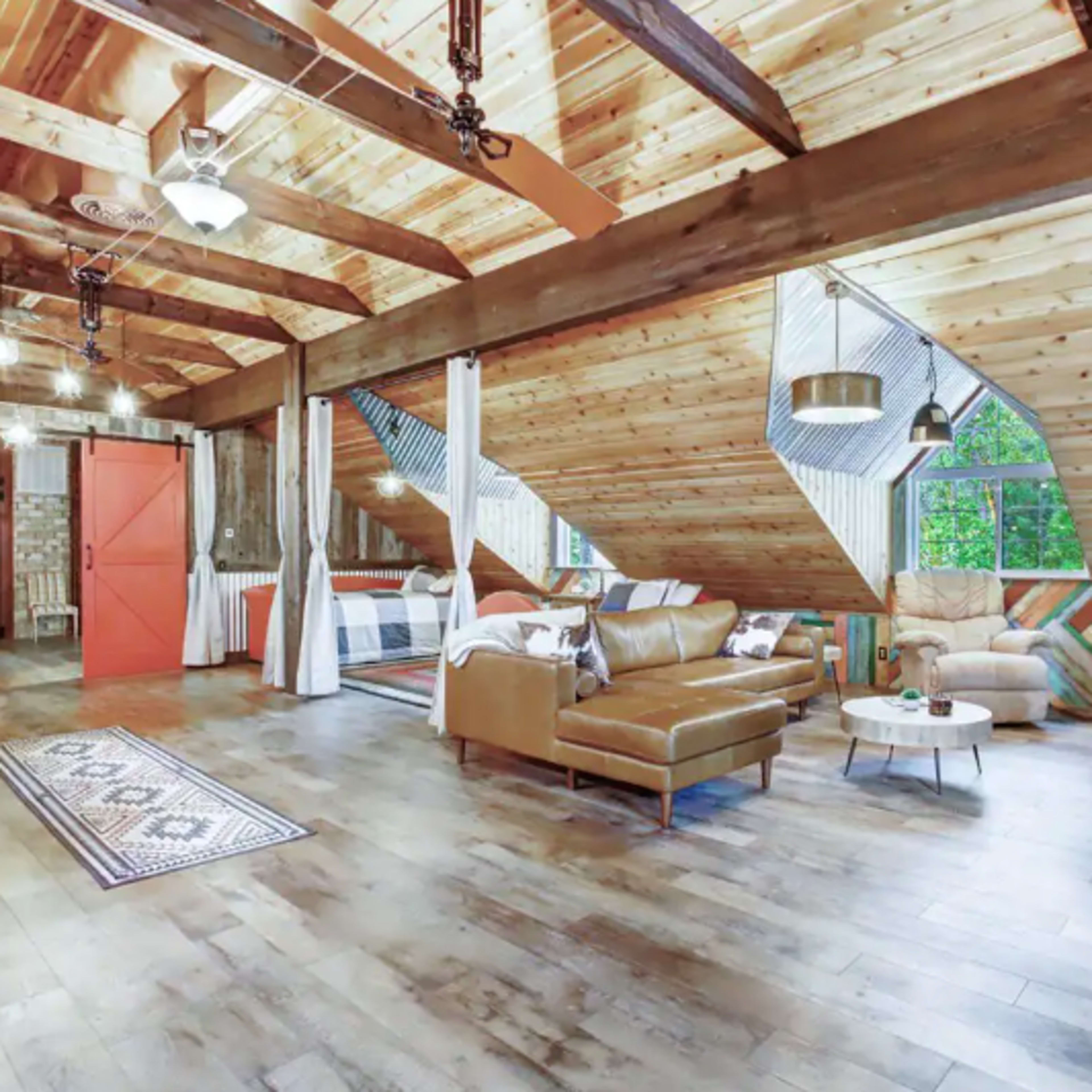 The room features a spacious, rustic interior with wooden beams, a leather sofa, and large windows that allow natural light to fill the space.