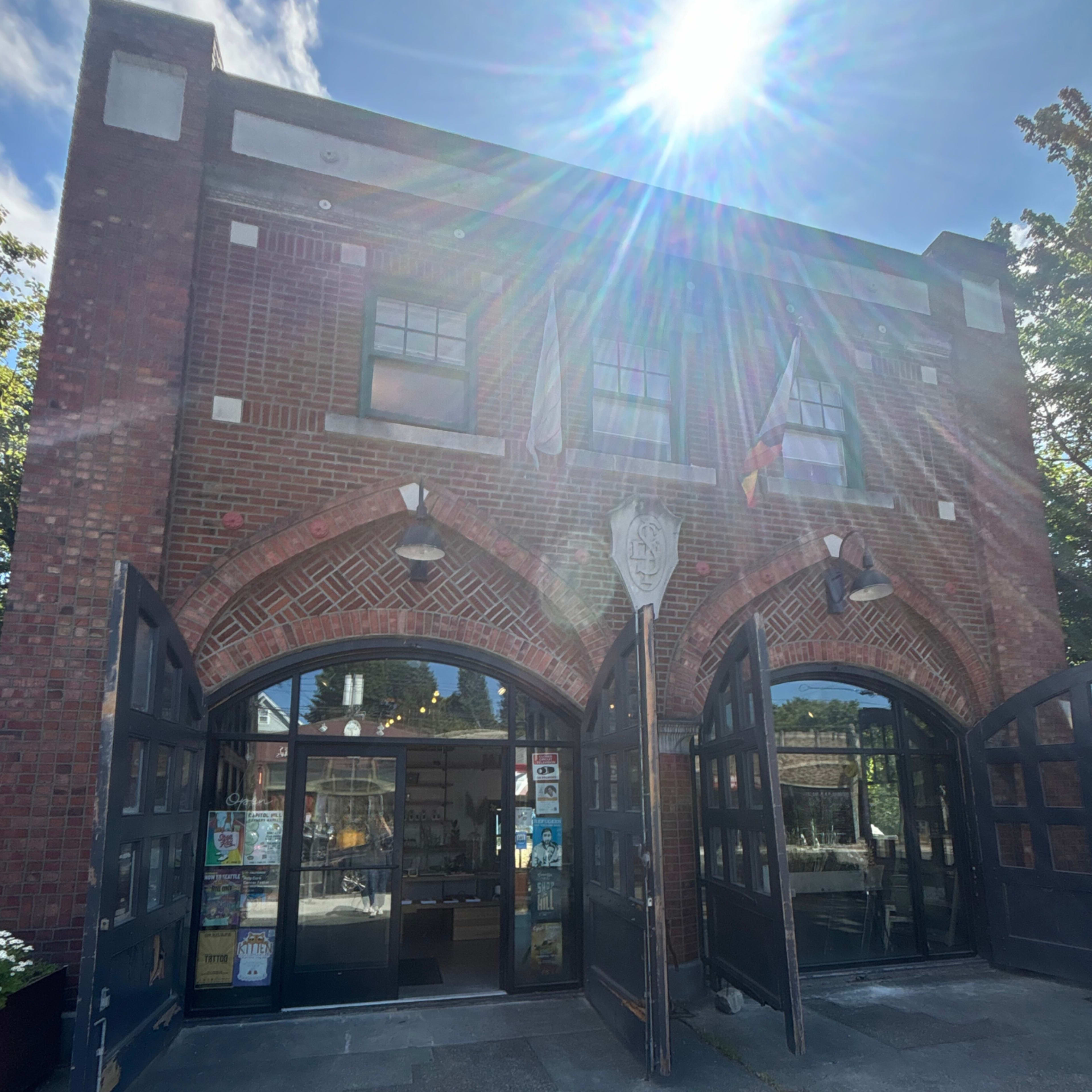 A brick building with arched double doors open to reveal a brightly lit interior, featuring several flags and a sunburst effect in the sky.
