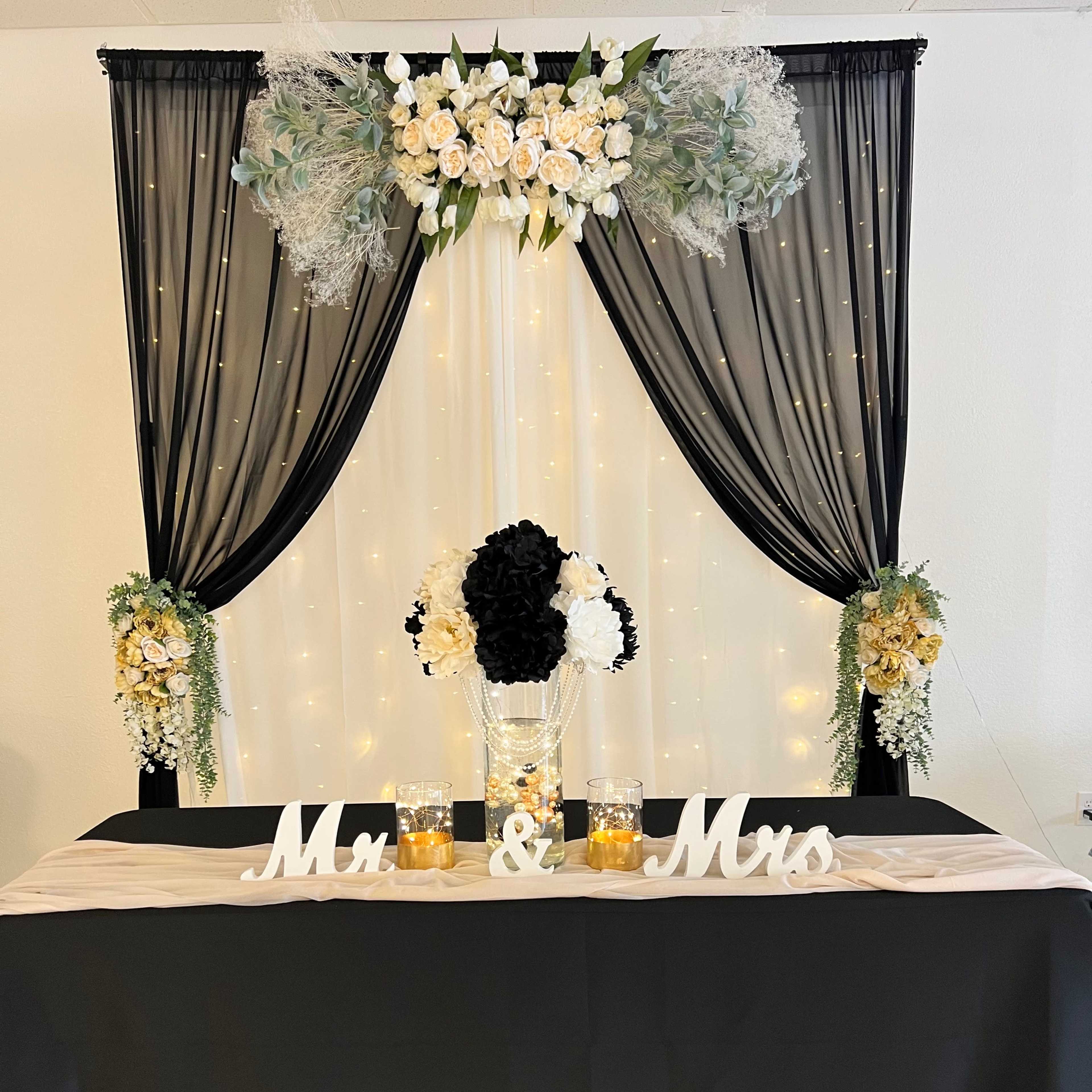 A decorated table with a black tablecloth features a floral arrangement, candles, and a "Mr. & Mrs." sign, set against a backdrop of flowing black and white drapery with fairy lights.