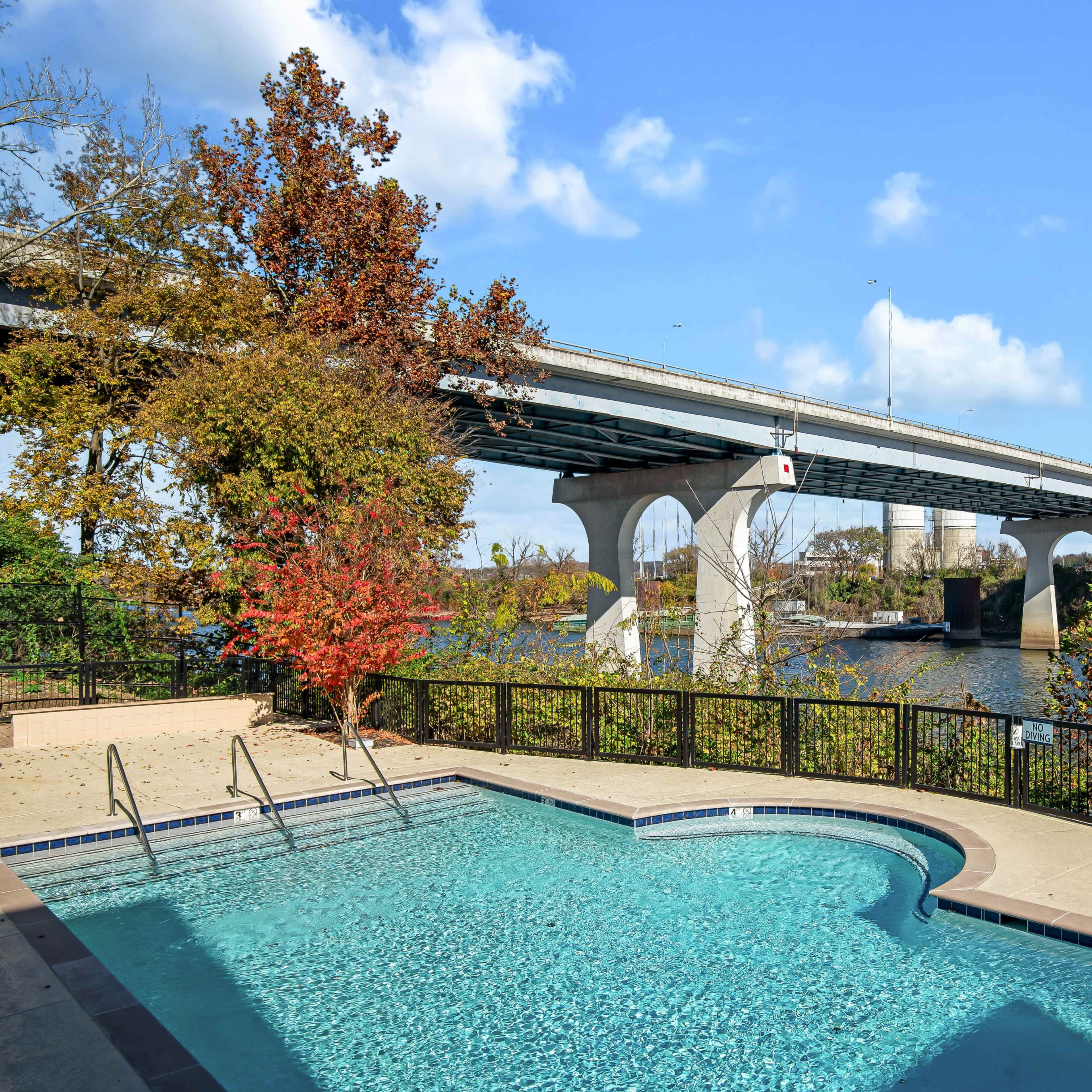 A swimming pool is located near a river, with a bridge spanning across the water in the background.