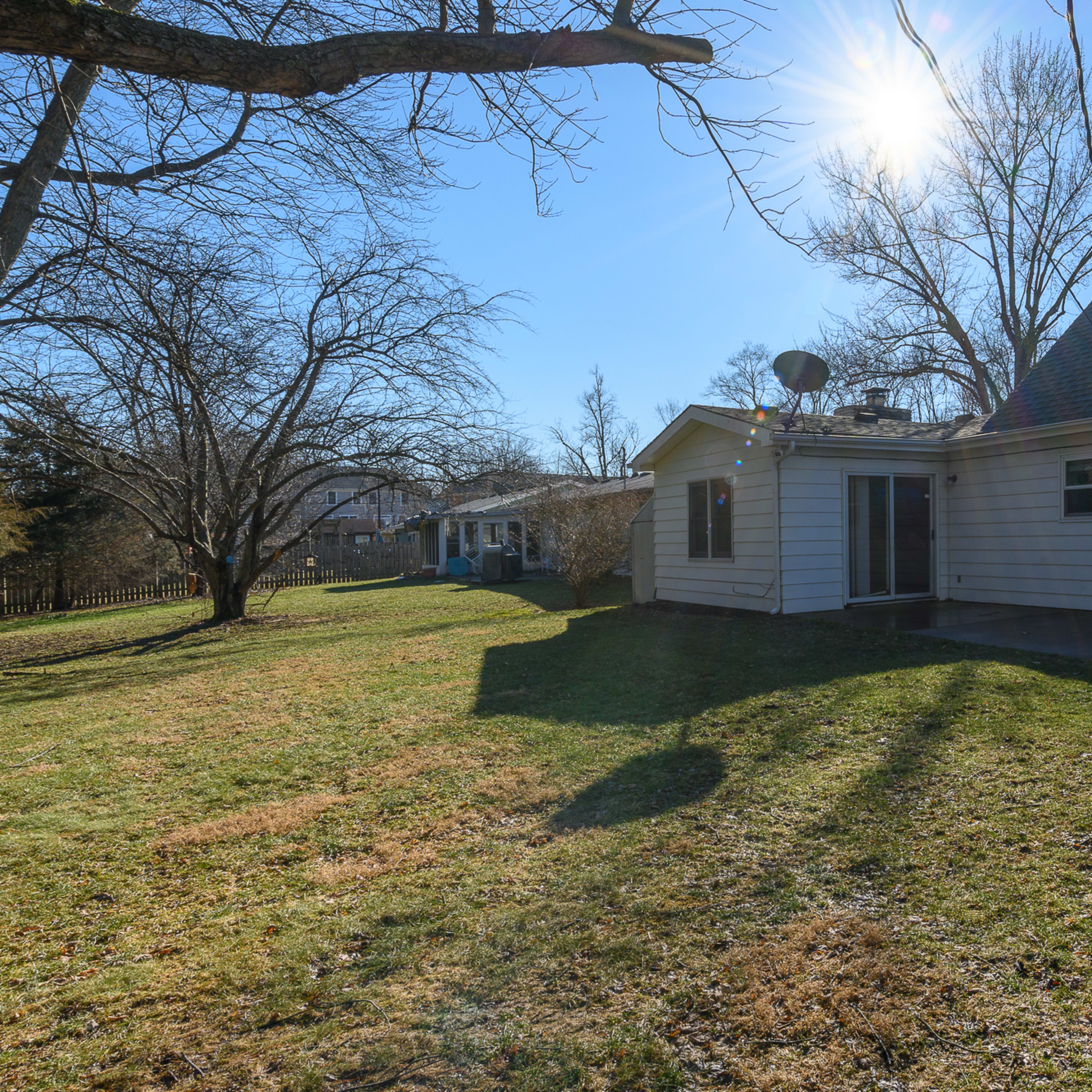 A house with a light-colored exterior is shown in a grassy backyard under a clear blue sky with a visible sun flare.