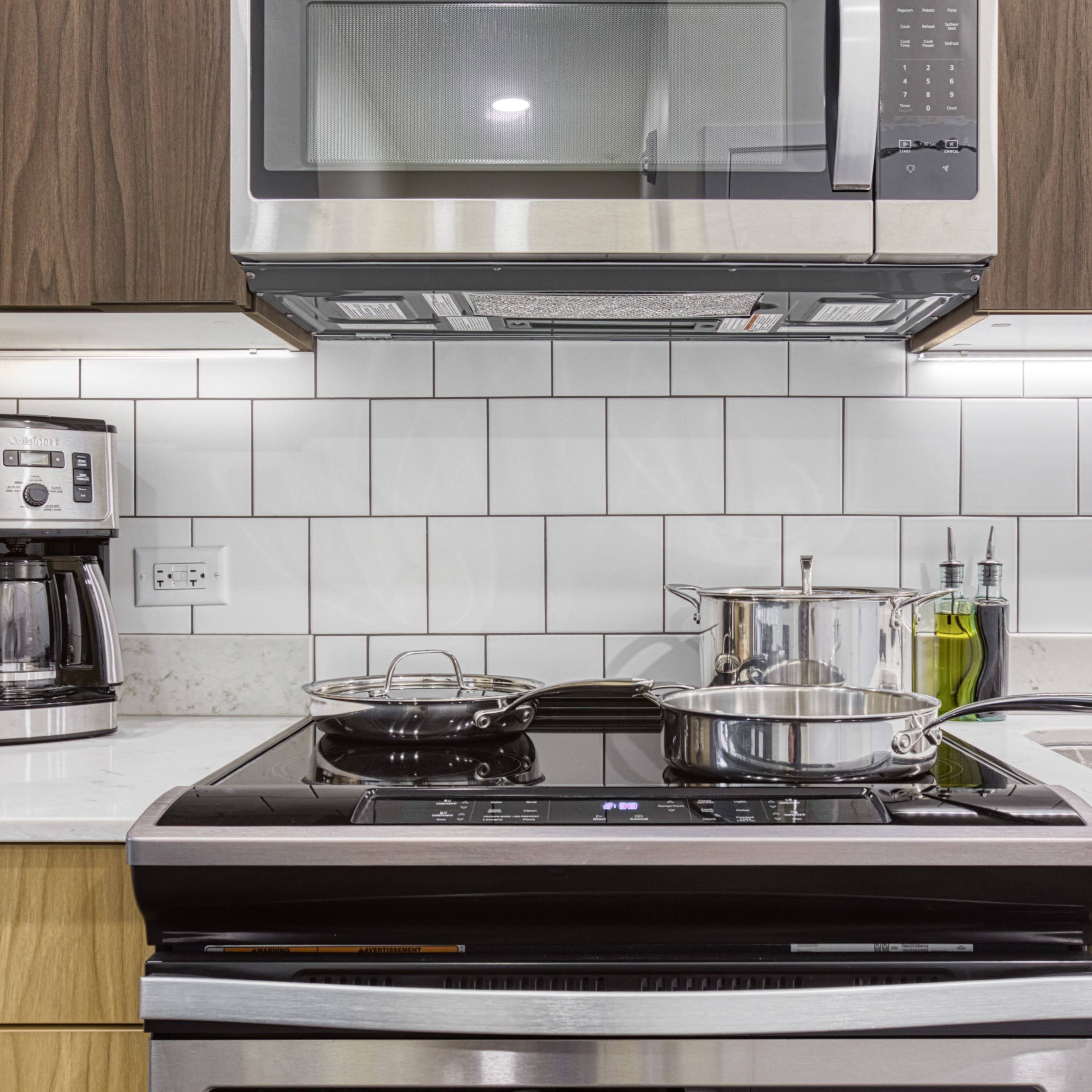 A modern kitchen countertop featuring a stove with pots, a microwave above, a coffee maker, and olive oil on the side.