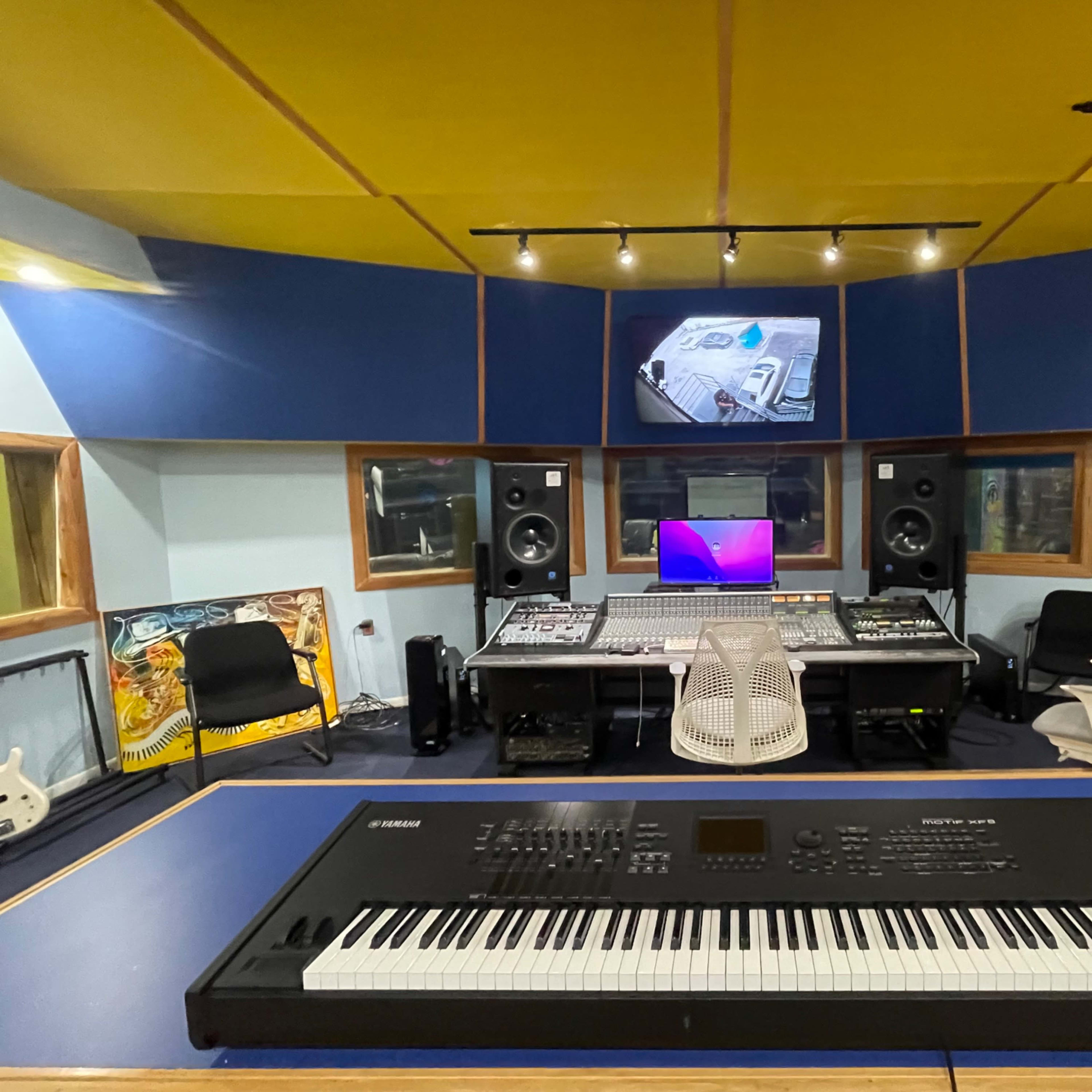 A recording studio with a digital keyboard in the foreground, a mixing console, speakers, and a computer monitor in the background.