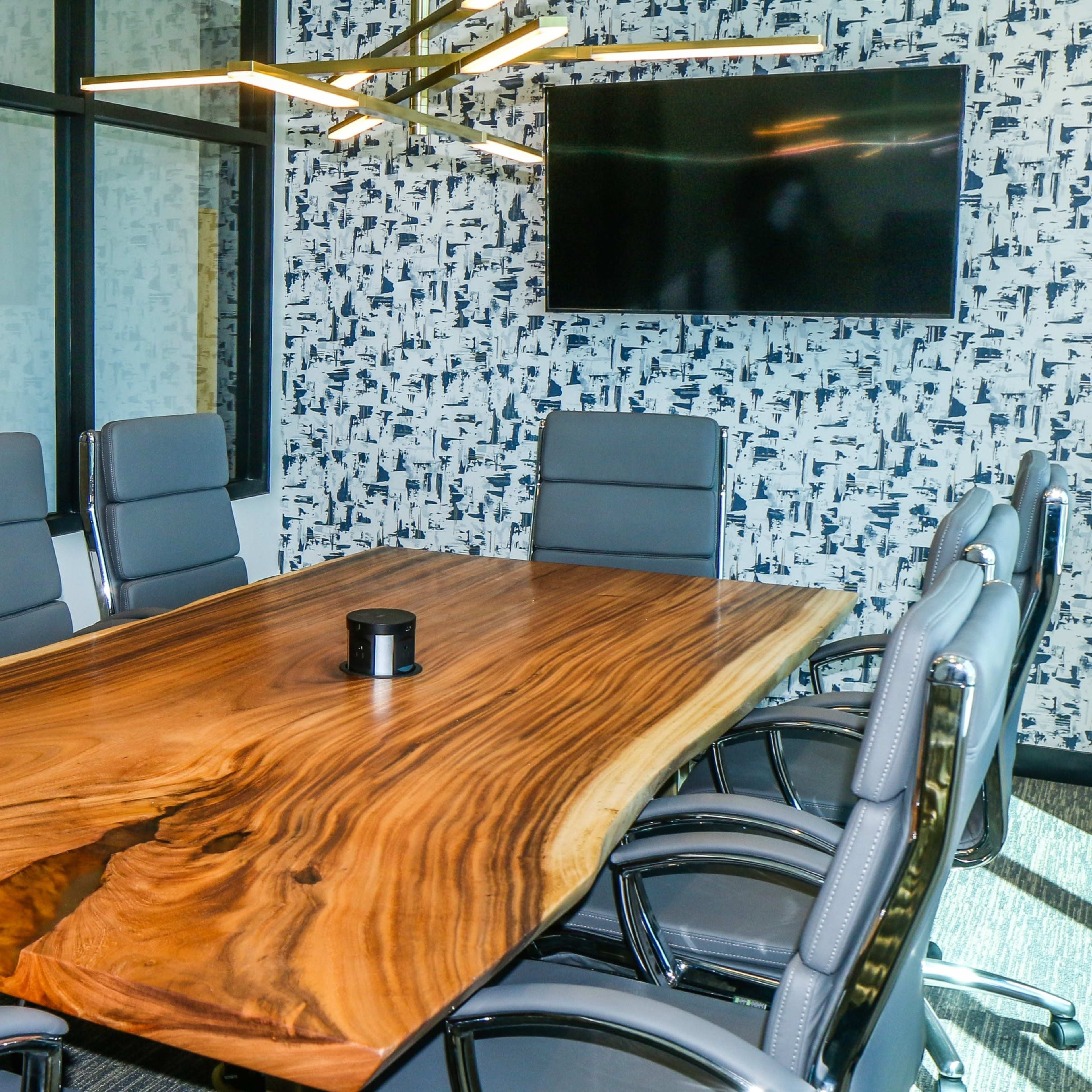 A conference room features a large wooden table surrounded by gray leather chairs, with a television and decorative wallpaper in the background.