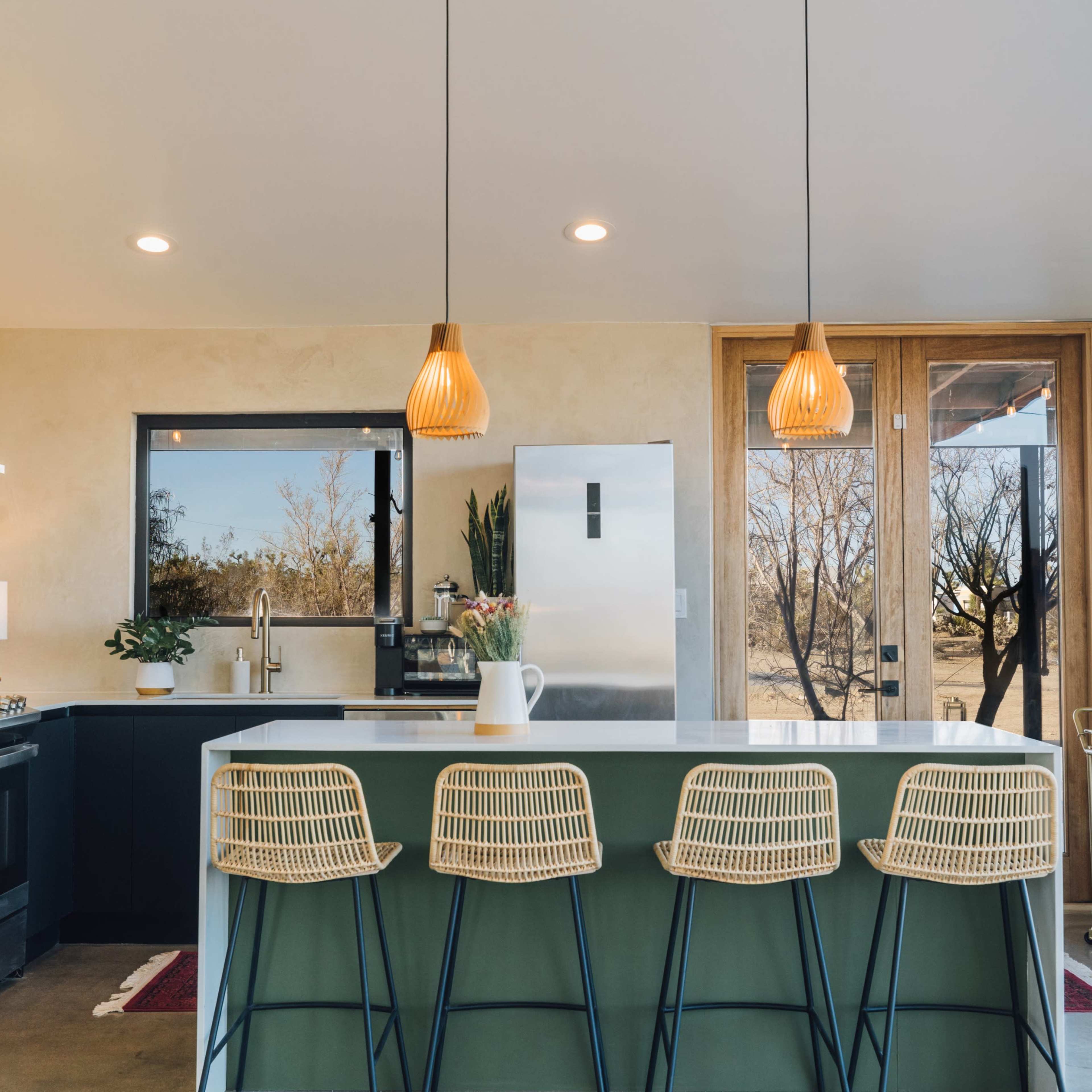 A modern kitchen features a large island with four bar stools, stainless steel appliances, and large windows providing a view of the outdoors.