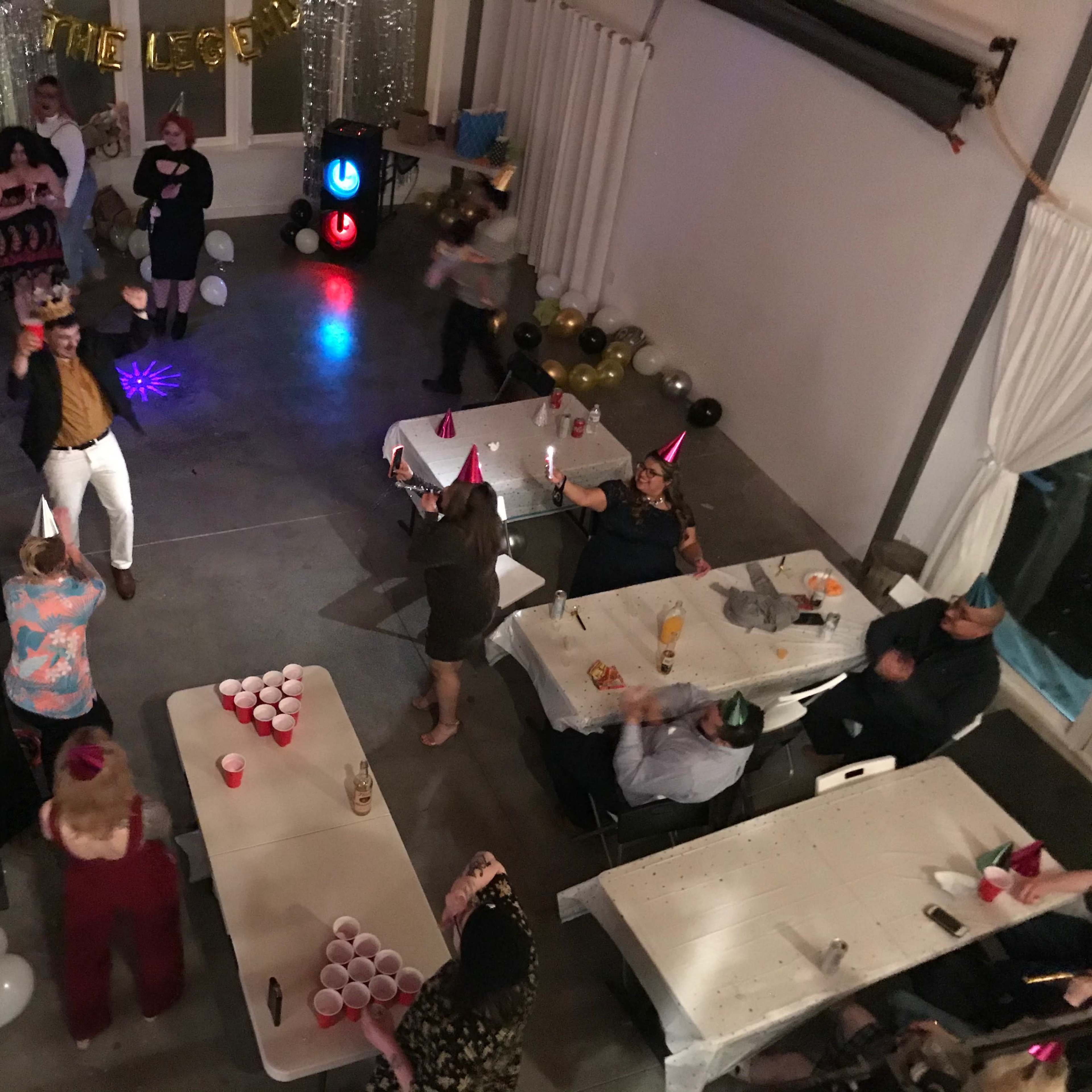 A crowded party scene shows guests dancing and socializing around tables adorned with drinks and decorations, while a colorful light illuminates the floor.